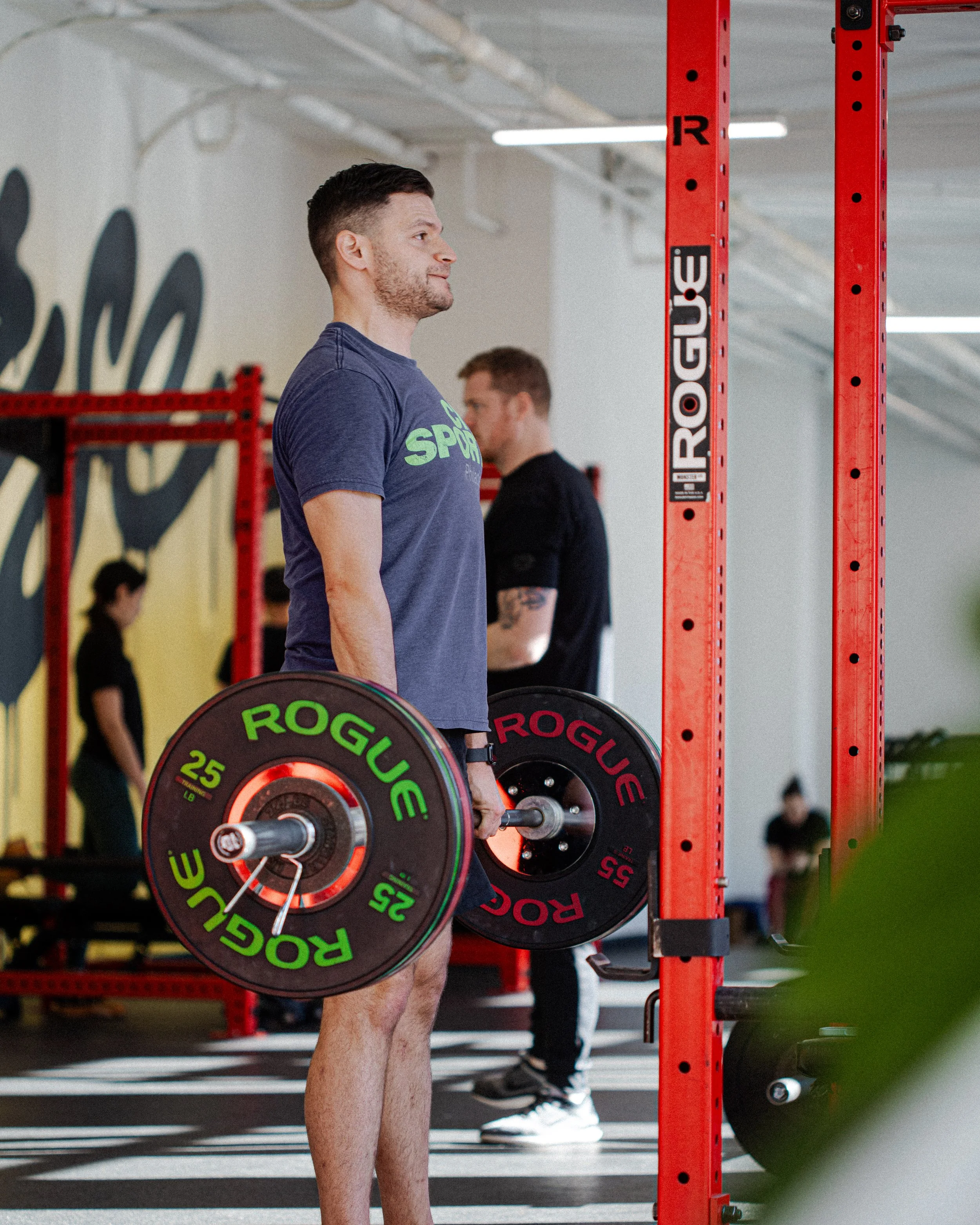 Man lifting weights in a gym, holding a loaded barbell, with others working out in background.