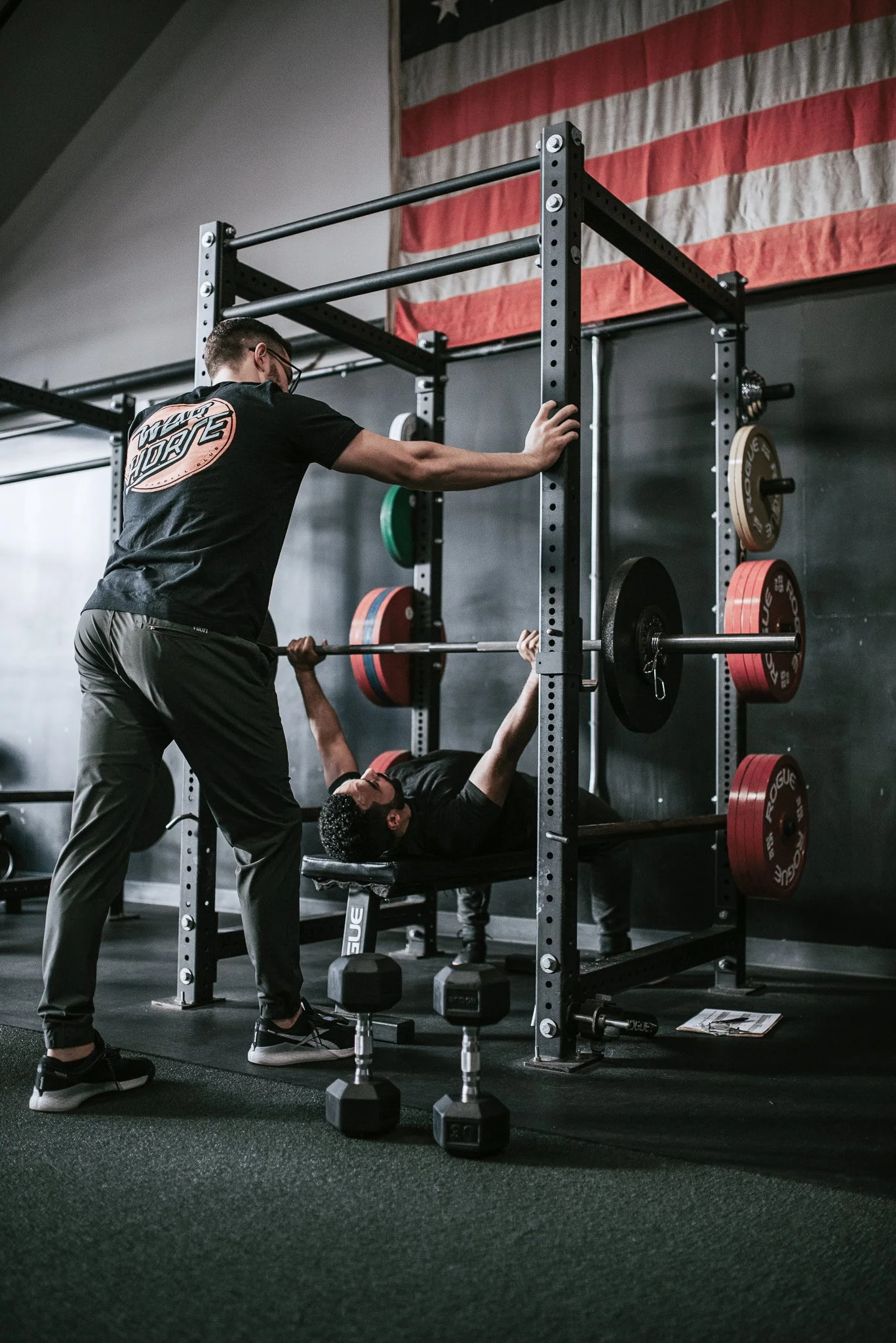 A person lying on a bench pressing a barbell with weight plates while a trainer spotlights them, in a gym with an American flag hanging on the wall.
