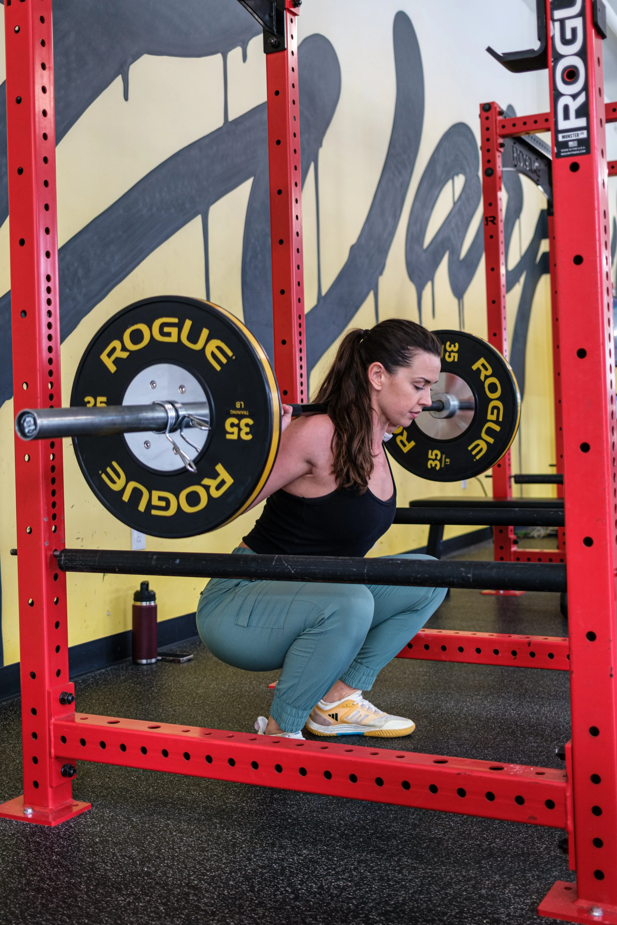 A woman with brown hair tied back, wearing a black tank top, teal pants, and white sneakers, doing a squat exercise with a barbell loaded with Rogues weight plates in a gym with yellow walls and red racks.