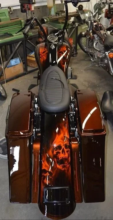 Motorcycle with a black and orange skull design painted on the gas tank and rear fender, parked indoors with other motorcycles nearby.