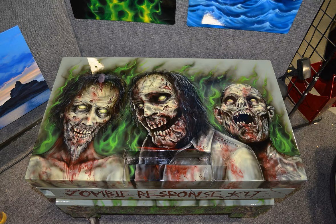 Airbrushed artwork of three zombie figures with green flames behind them, displayed on a box labeled 'Zombie Response Kit' at an art exhibit.