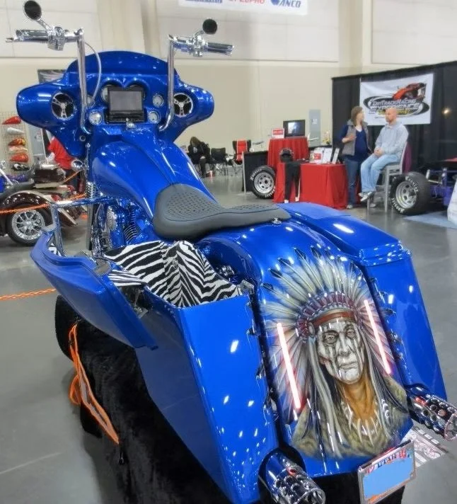 A custom blue motorcycle displayed at an indoor exhibition. It features artwork of a Native American chief wearing a feathered headdress on the front panel, and zebra-patterned cloth on the seat.