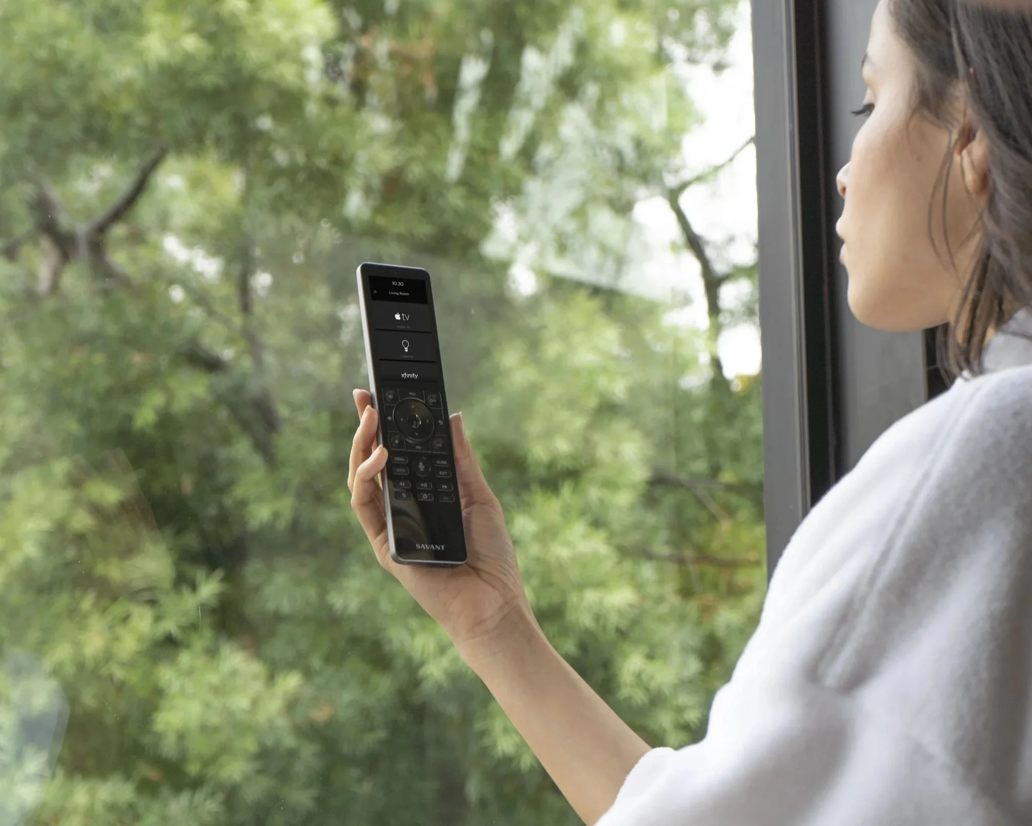 Woman using a remote control to manage connected home technology, with outdoor views visible through the window