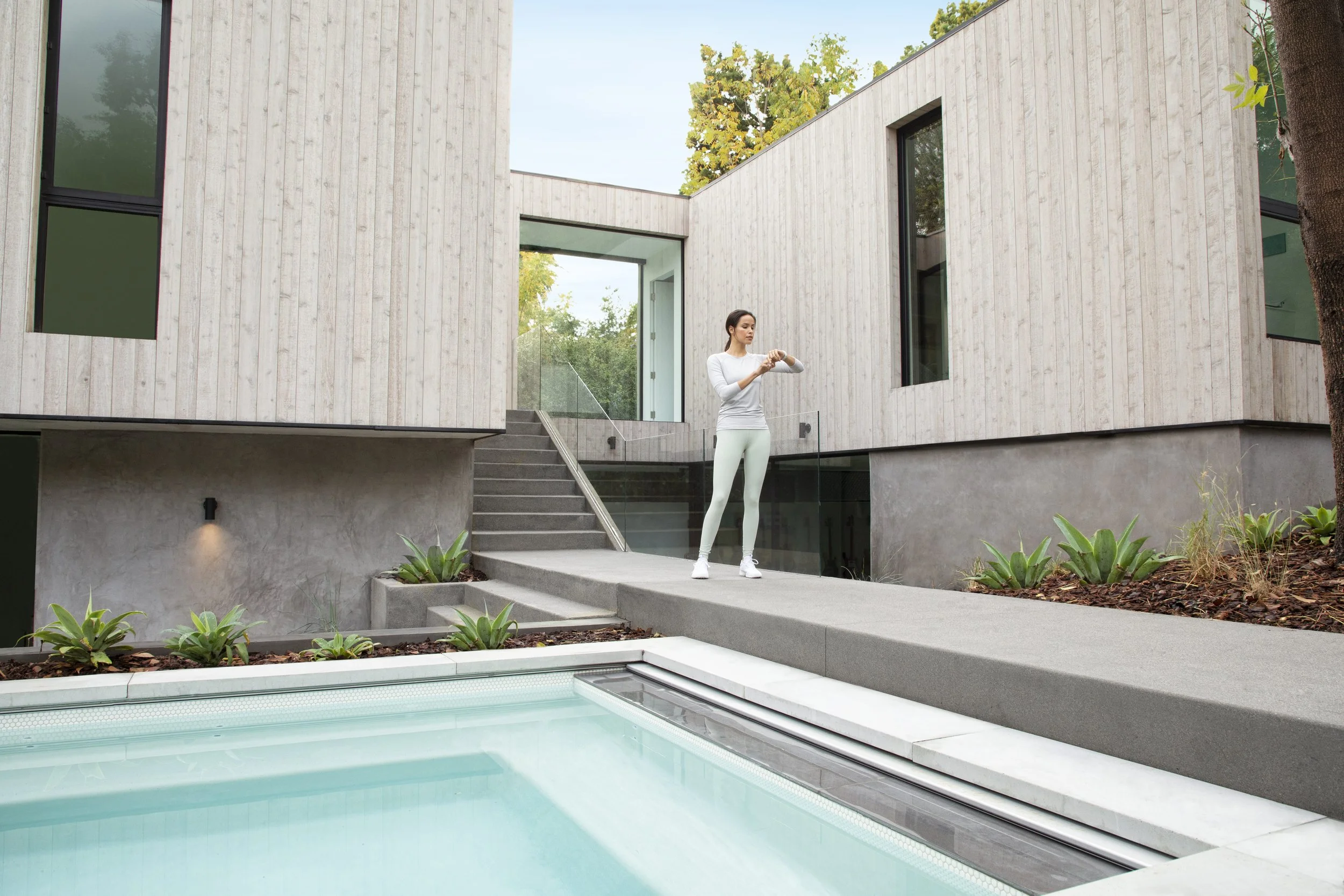 A woman standing on a concrete patio beside a swimming pool, checking her watch outside a modern house with large windows and wooden siding.