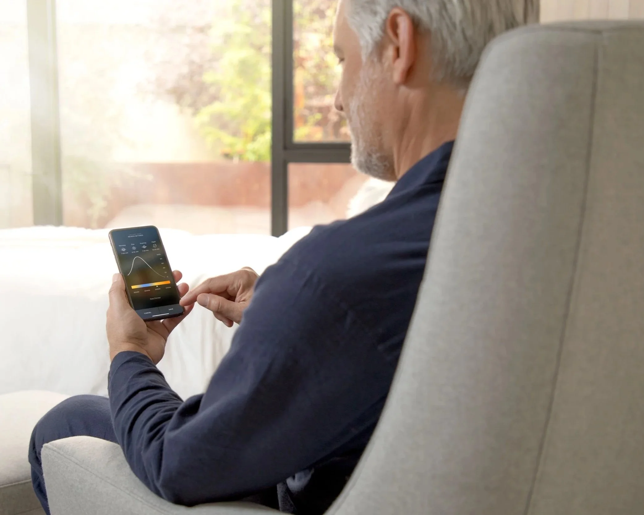 Man using a smartphone to monitor lighting and climate controls in a modern room with garden views
