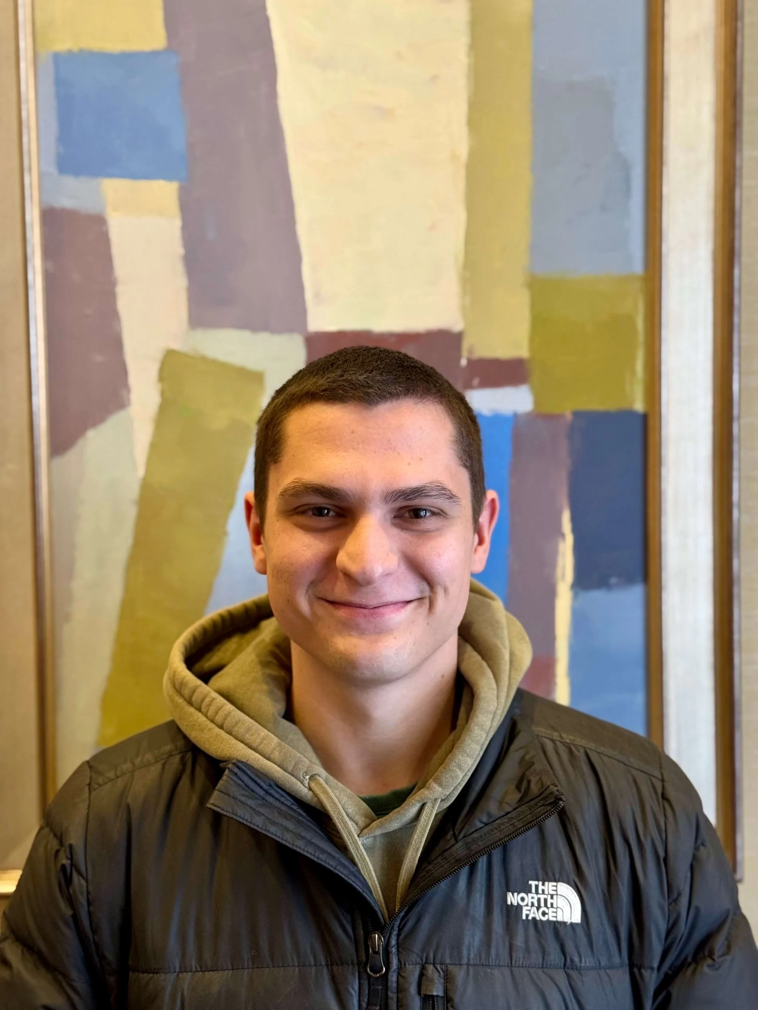 A young man with short dark hair, smiling, wearing a beige hoodie and a black The North Face jacket, standing in front of an abstract painting with blocks of pastel and earth tones.