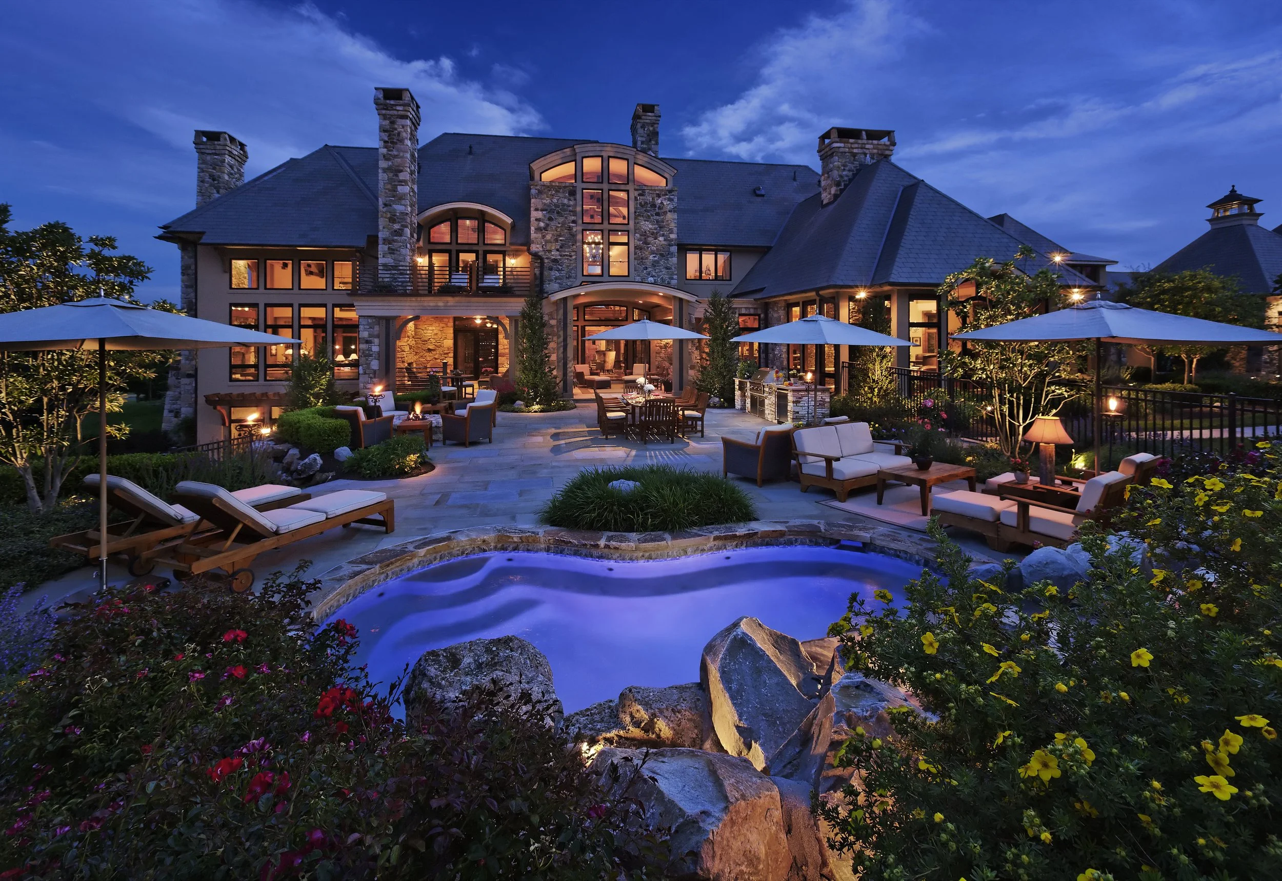 Luxury house with stone and wood exterior, illuminated windows, outdoor seating area with umbrellas, lounge chairs, and a hot tub in the garden during evening.
