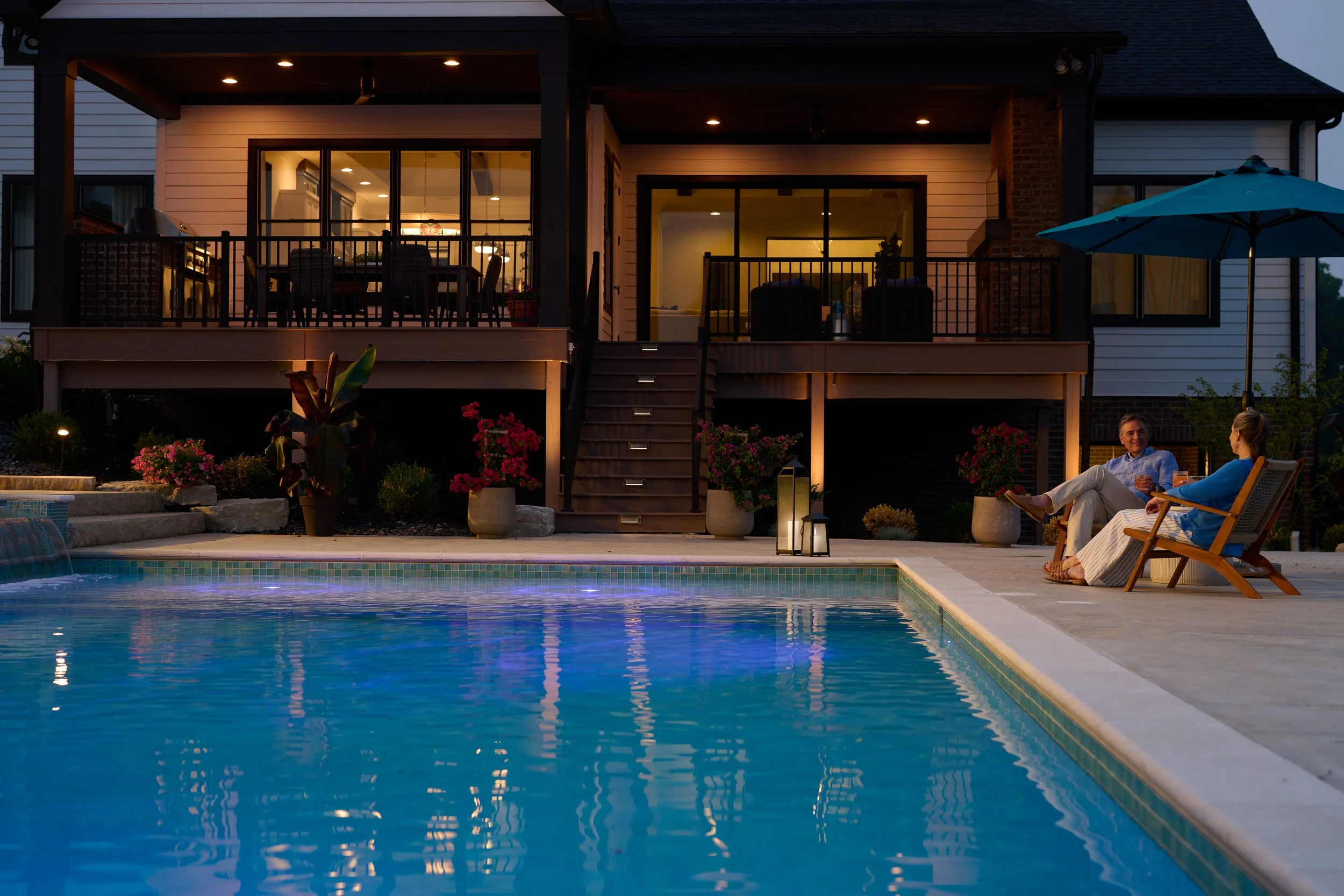 A backyard at dusk with a lit swimming pool, potted plants, and two people sitting in wooden lounge chairs under an umbrella, enjoying drinks.
