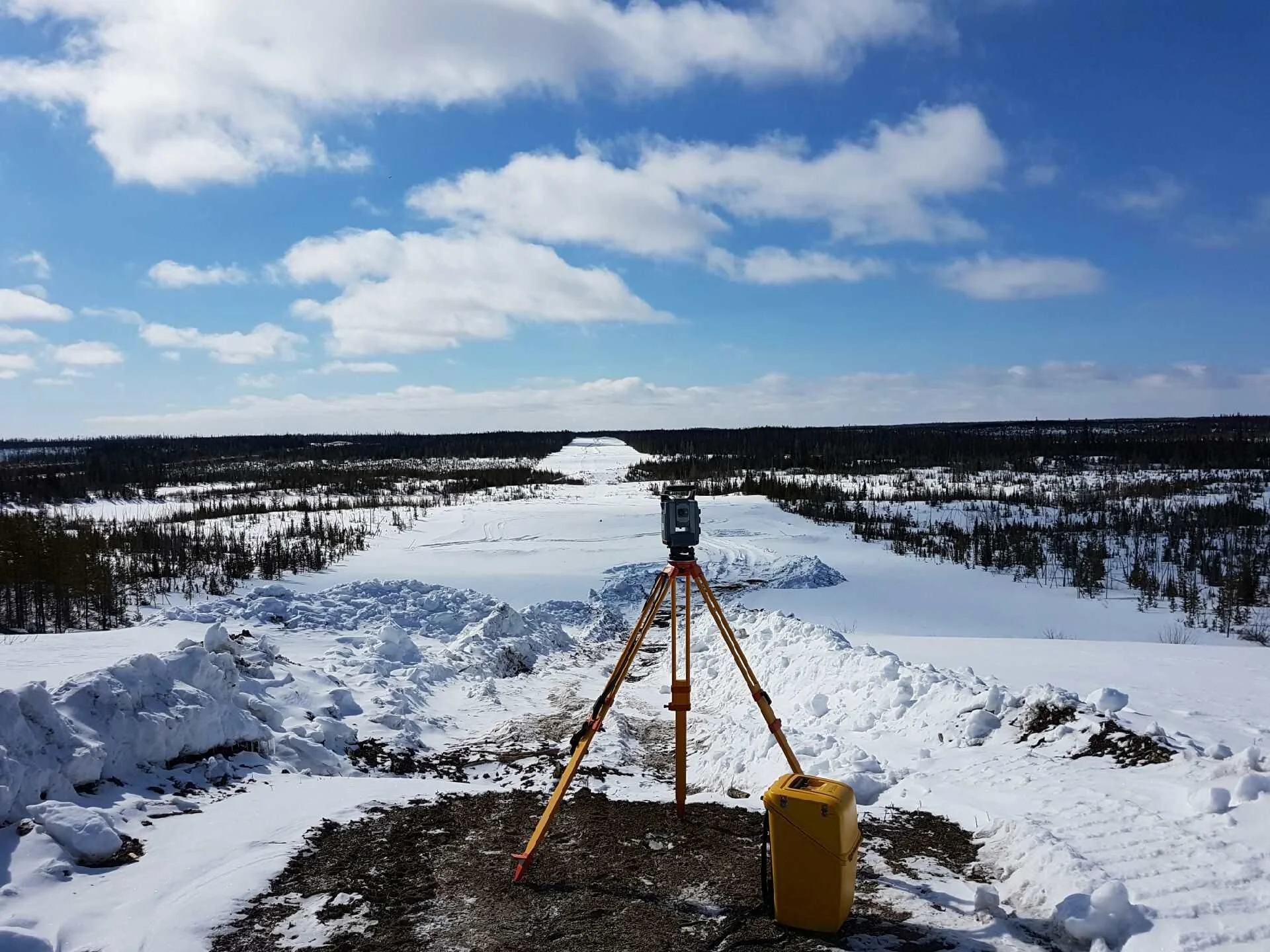 A total station mouted on a yellow tripad stands on a snow-covered embankment overlooking a vast, frozen lanscape with scattered trees. Fresh snowbanks line the foreground, and a bright blue sky with scattered clouds stretches overhead.