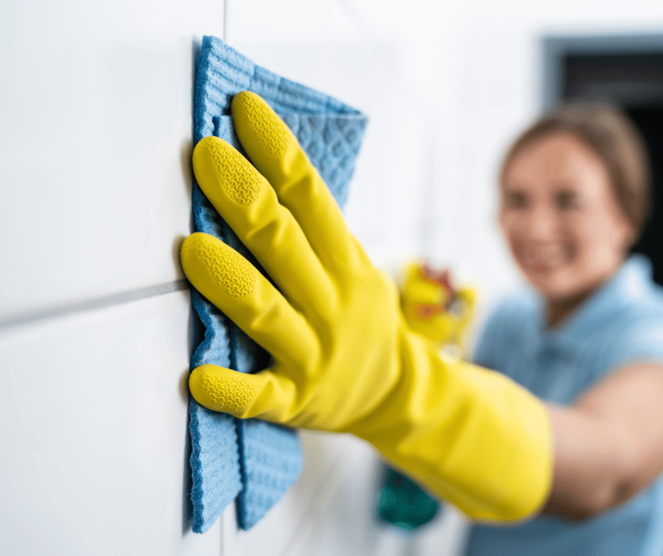 Person wearing yellow rubber gloves and cleaning a white tiled wall with a blue cleaning cloth, smiling in the background.