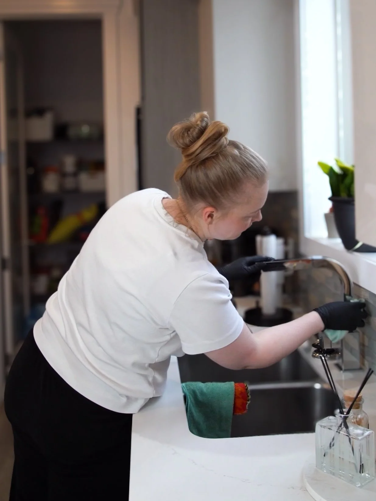 A woman with a bun hairstyle wearing a white t-shirt and black gloves cleaning a kitchen sink.