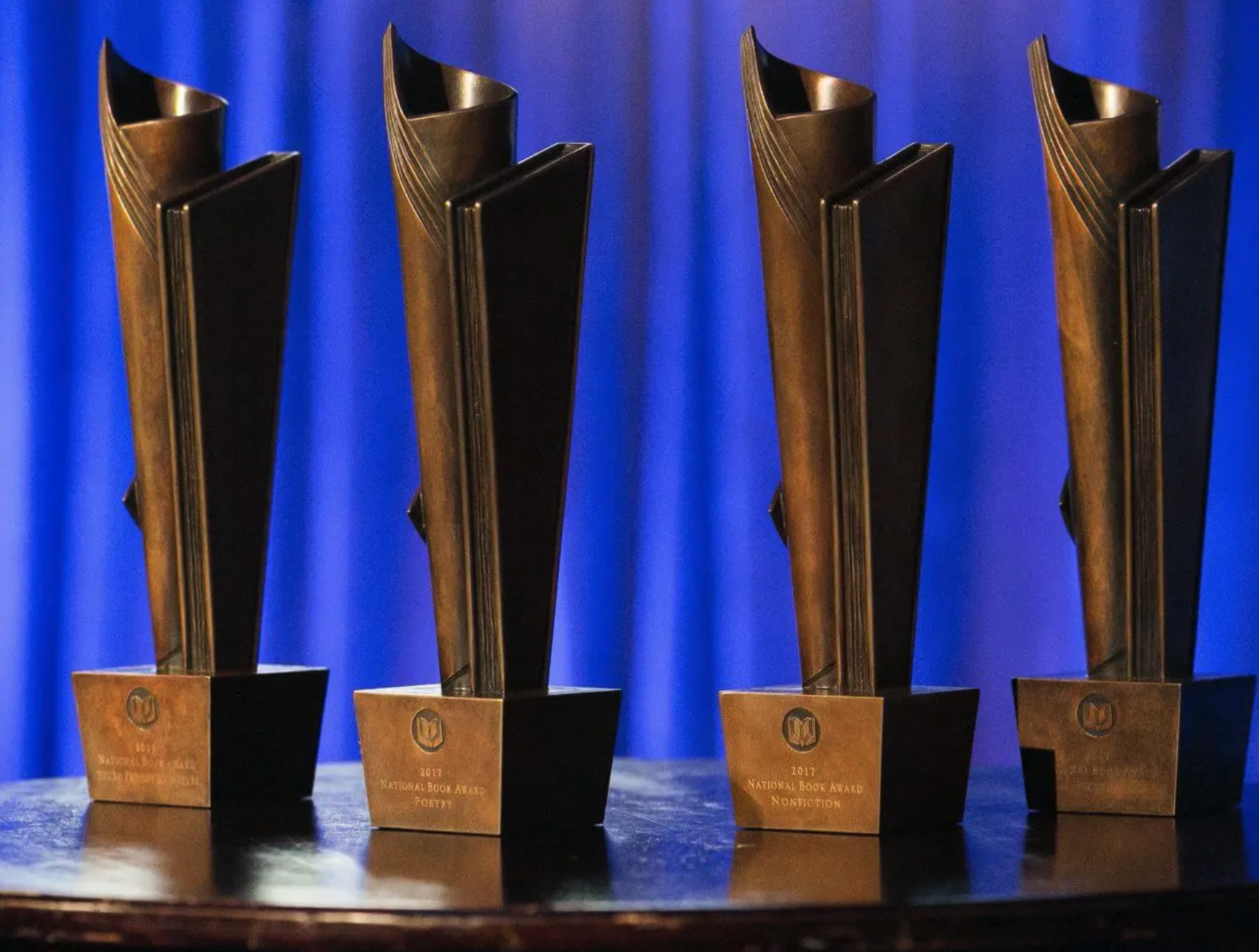 Four identical awards with a metallic gold and black design placed on a wooden surface, with a blue curtain background.