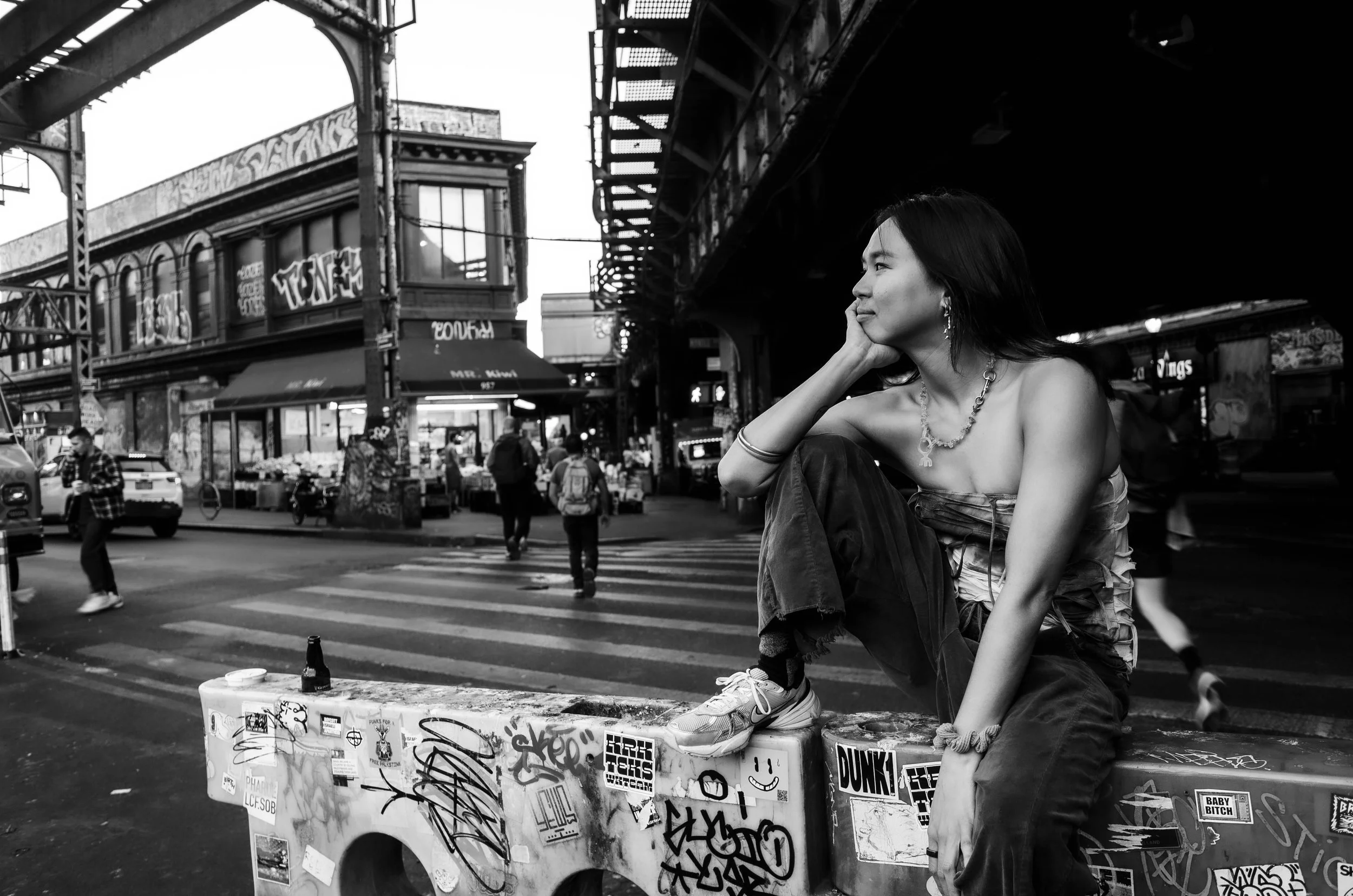 A photograph of Michelle Lhooq. Lhooq is sitting on a graffiti-covered surface with her leg resting on it under a bridge in an urban area, with pedestrians crossing the street behind her.