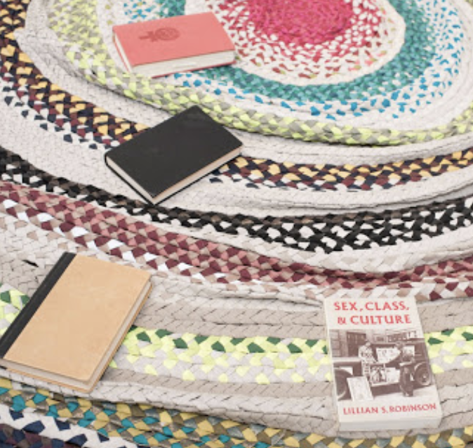 Colorful braided rugs, a pink book, a black notebook, a tan notebook, and a book titled 'Sex, Class, & Culture' on a display of woven rugs.