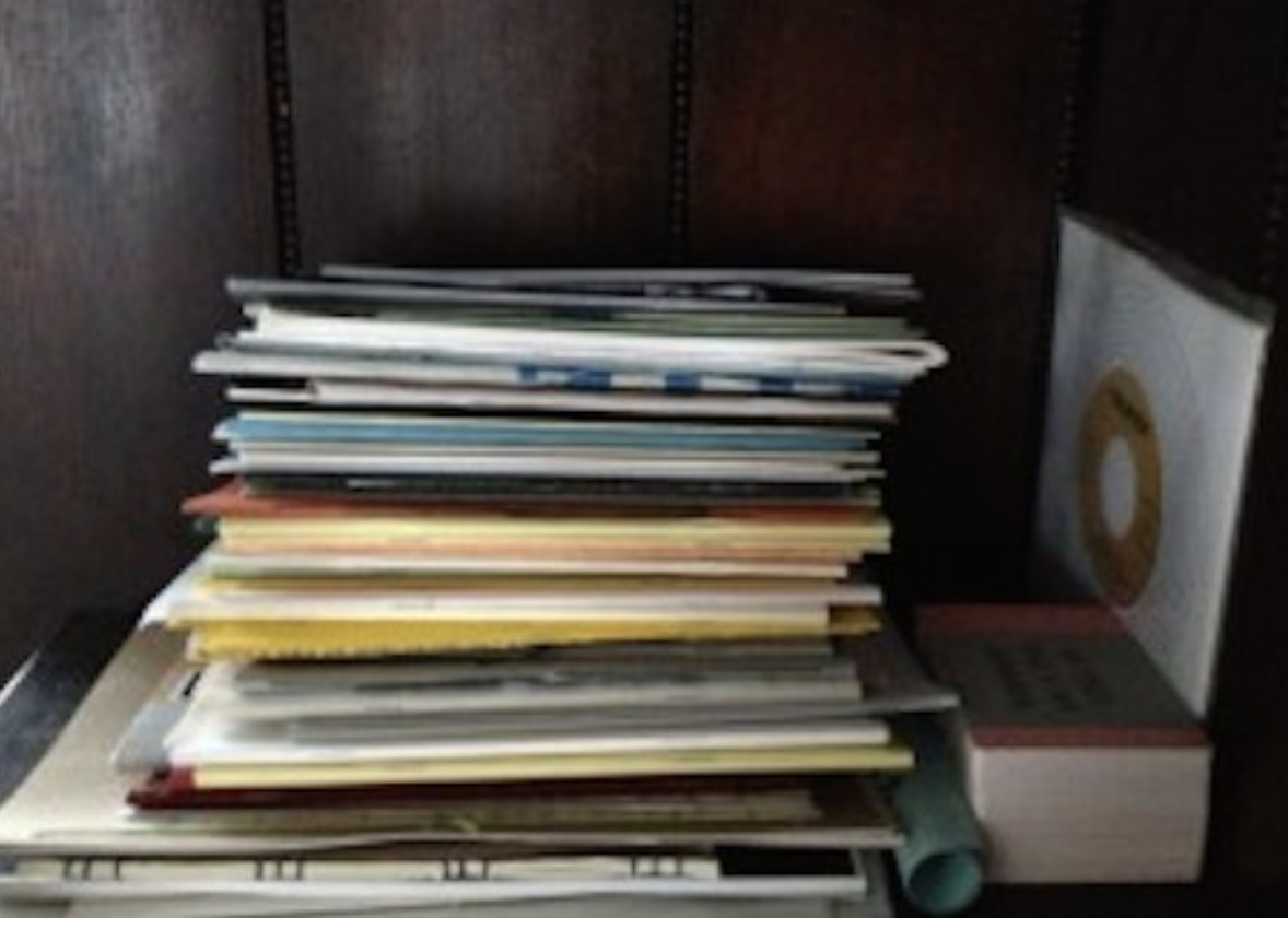 A bookshelf with a stack of magazines, books, and a paper sleeve with a CD on a wooden shelf.