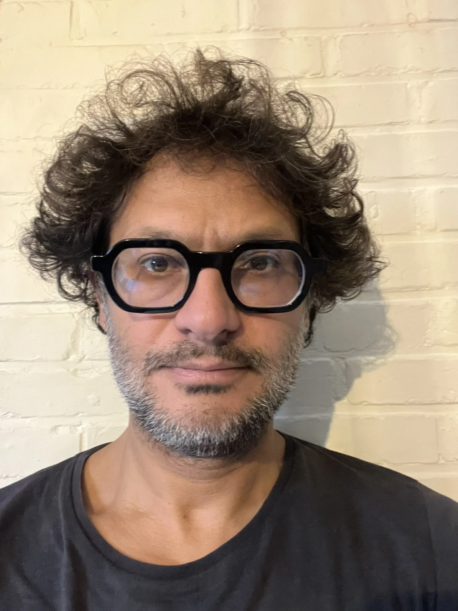 A photograph of Arun Saldanha. Saldanha has curly hair,  a short beard, and is wearing large black glasses. He is standing in front of a white brick wall.