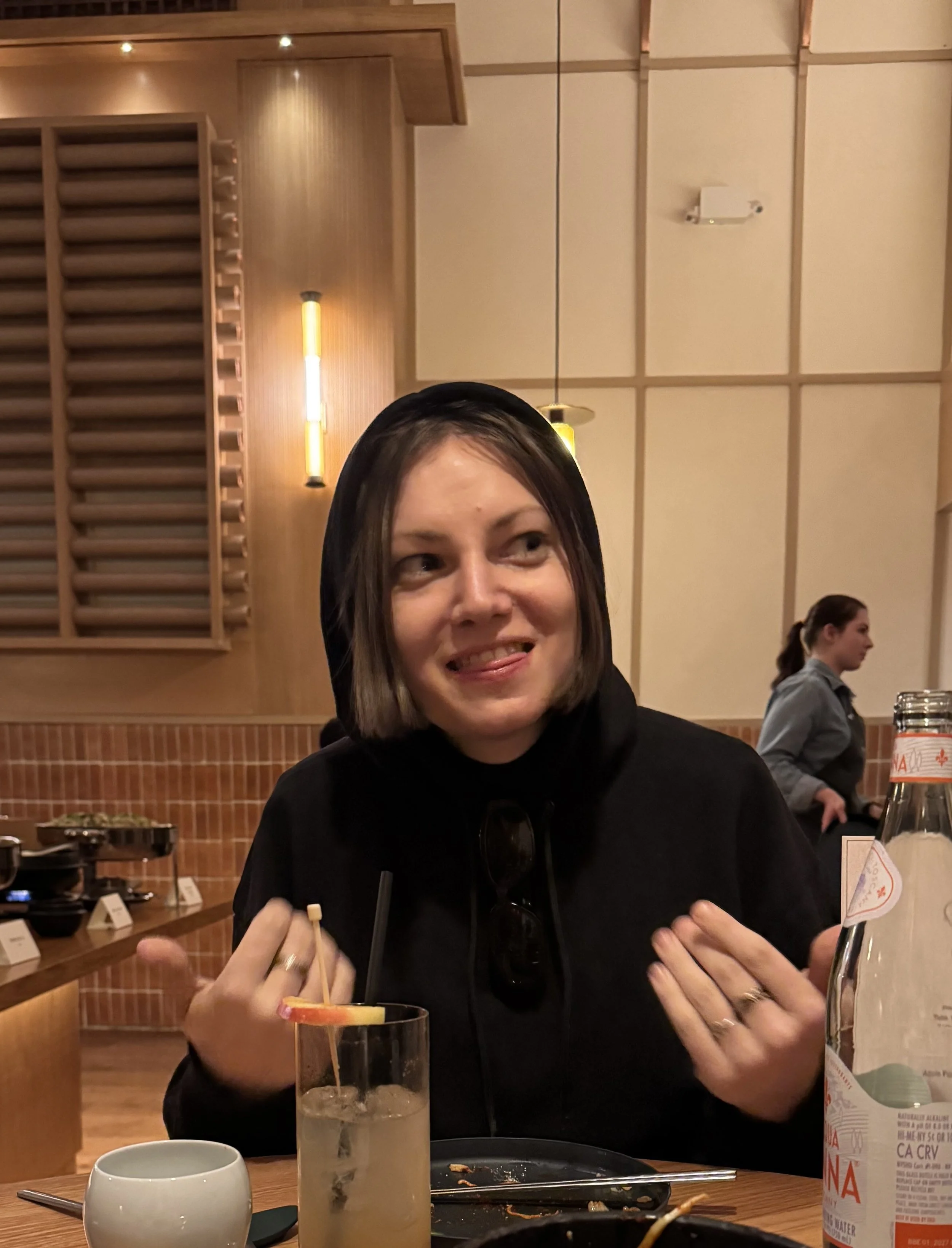 A photograph of hannah baer. baer has short brown hair and wears a dark hoodie. She is sitting at a restaurant table, making a playful face with tongue slightly out, with a drink and utensils on the table behind her.
