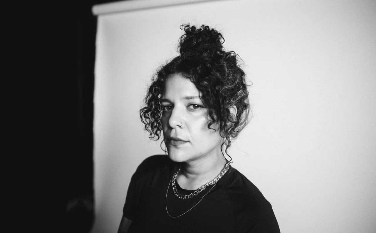A photograph of Sofía Córdova. Cordova has dark curly hair pinned up above her head and is sitting in front of a plain wall. She is looking into the camera seriously. 