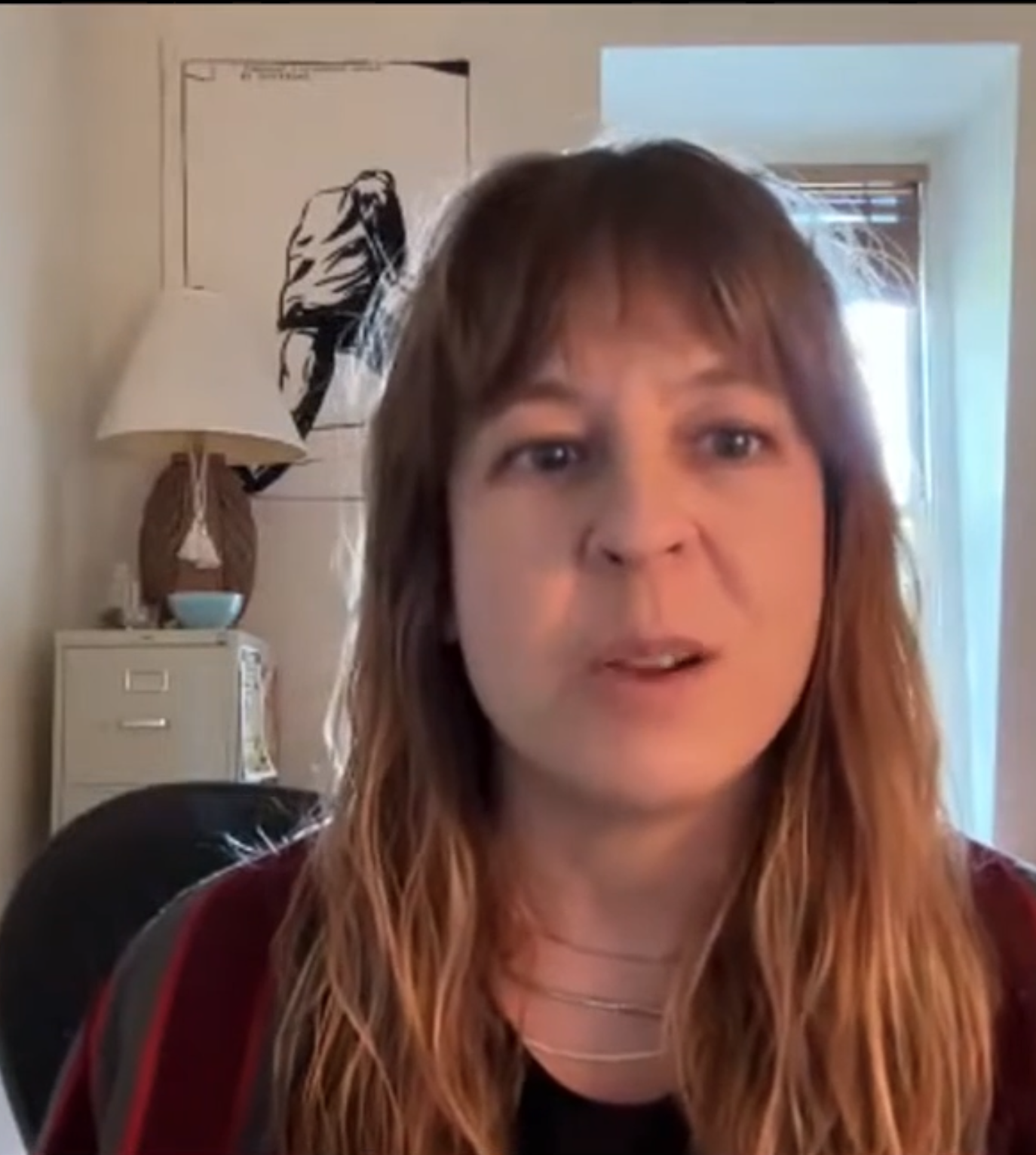 Woman with wavy, shoulder-length hair speaking during a video call in a room with a lamp, artwork, a window with blinds, and a white cabinet in the background.