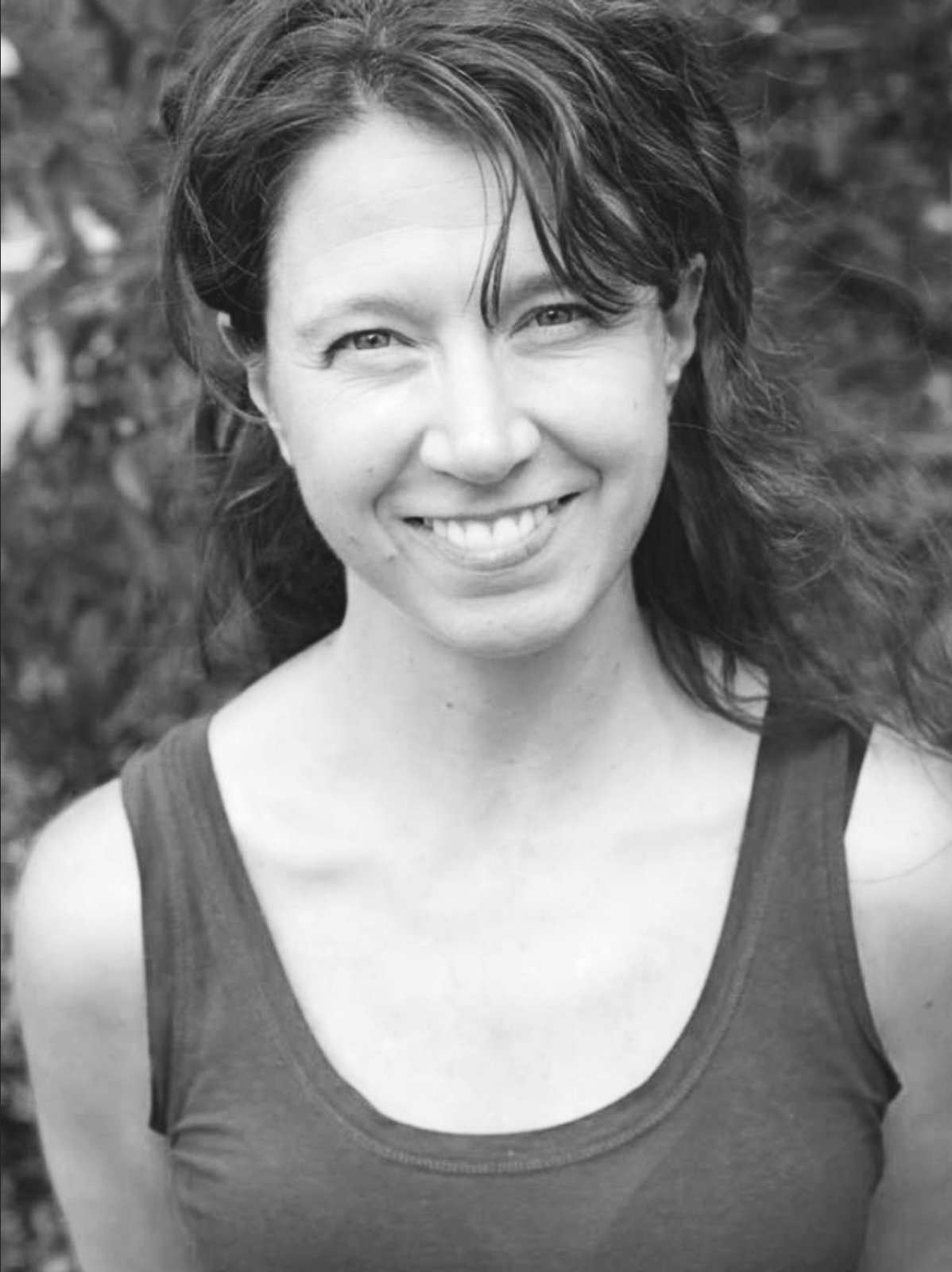 Black and white portrait of a smiling woman with short, wavy hair, wearing a sleeveless top, outdoors with blurred foliage in the background.