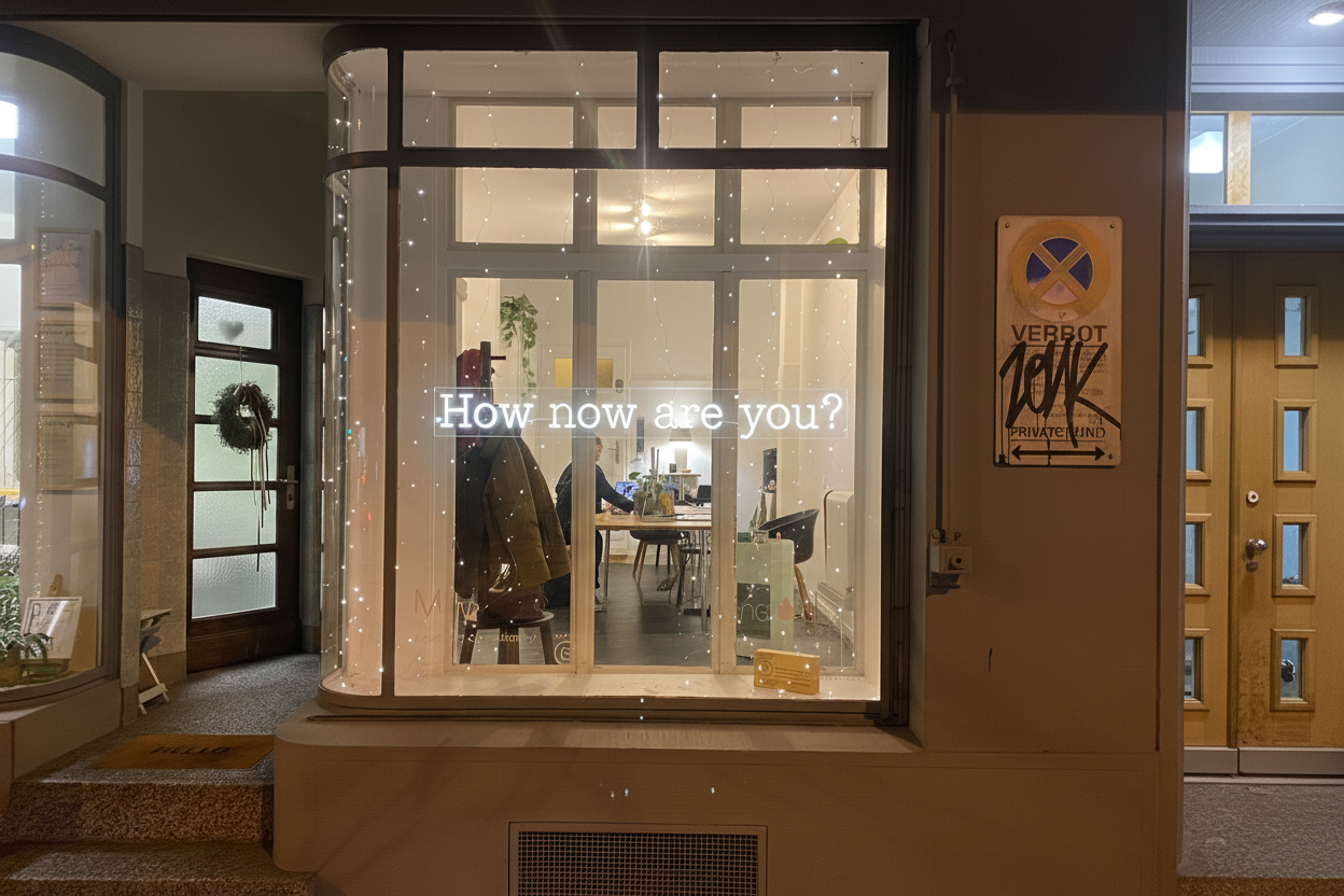 The window display shows the phrase "How now are you?" illuminated in white, with a view into a cozy, well-lit interior space where people are sitting at tables. There are string lights hanging around the window, and a coat hangs on a hook inside.