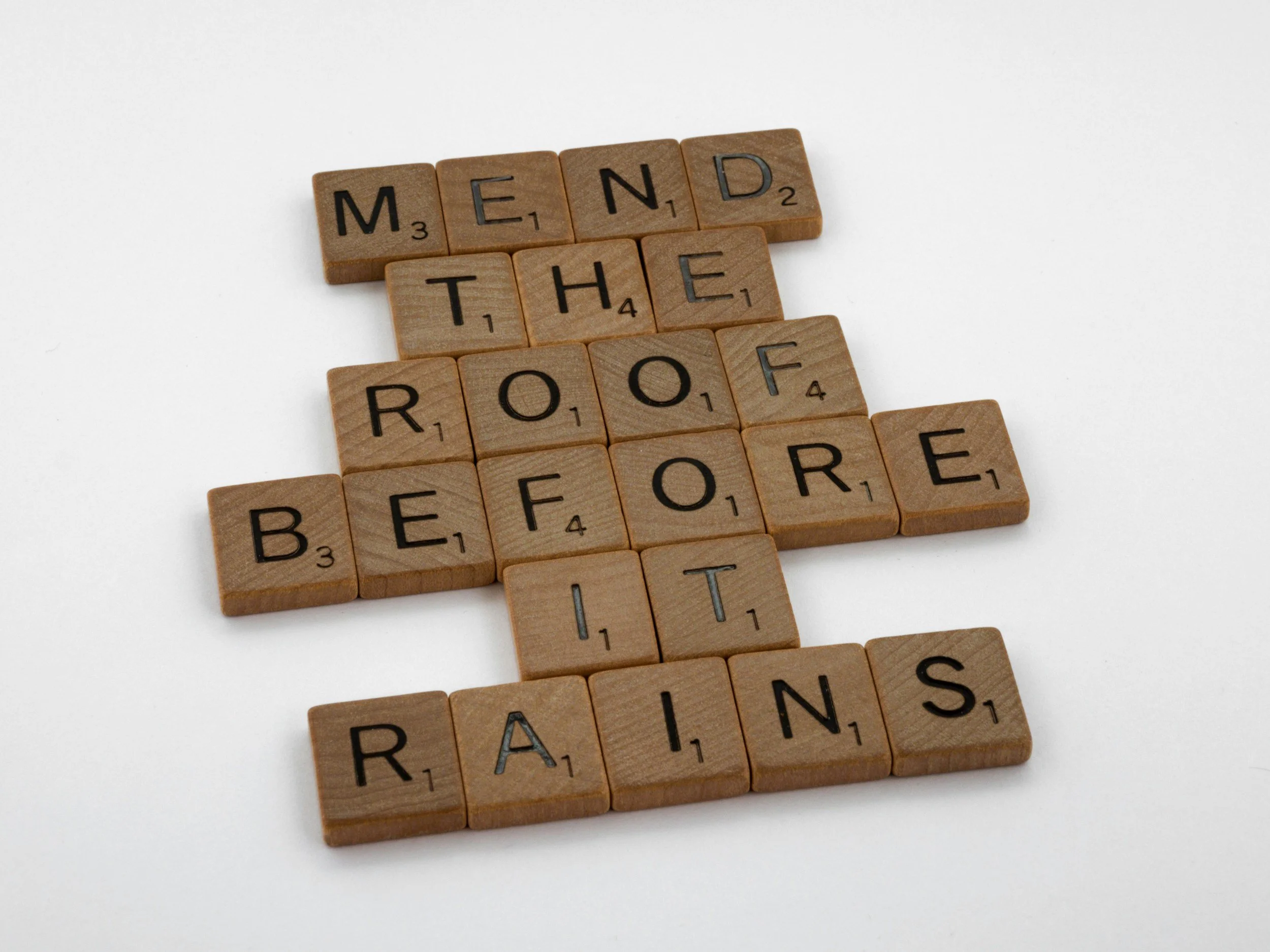 Scrabble tiles arranged to spell out 'MEND THE ROOF BEFORE IT RAINS' on a white background.
