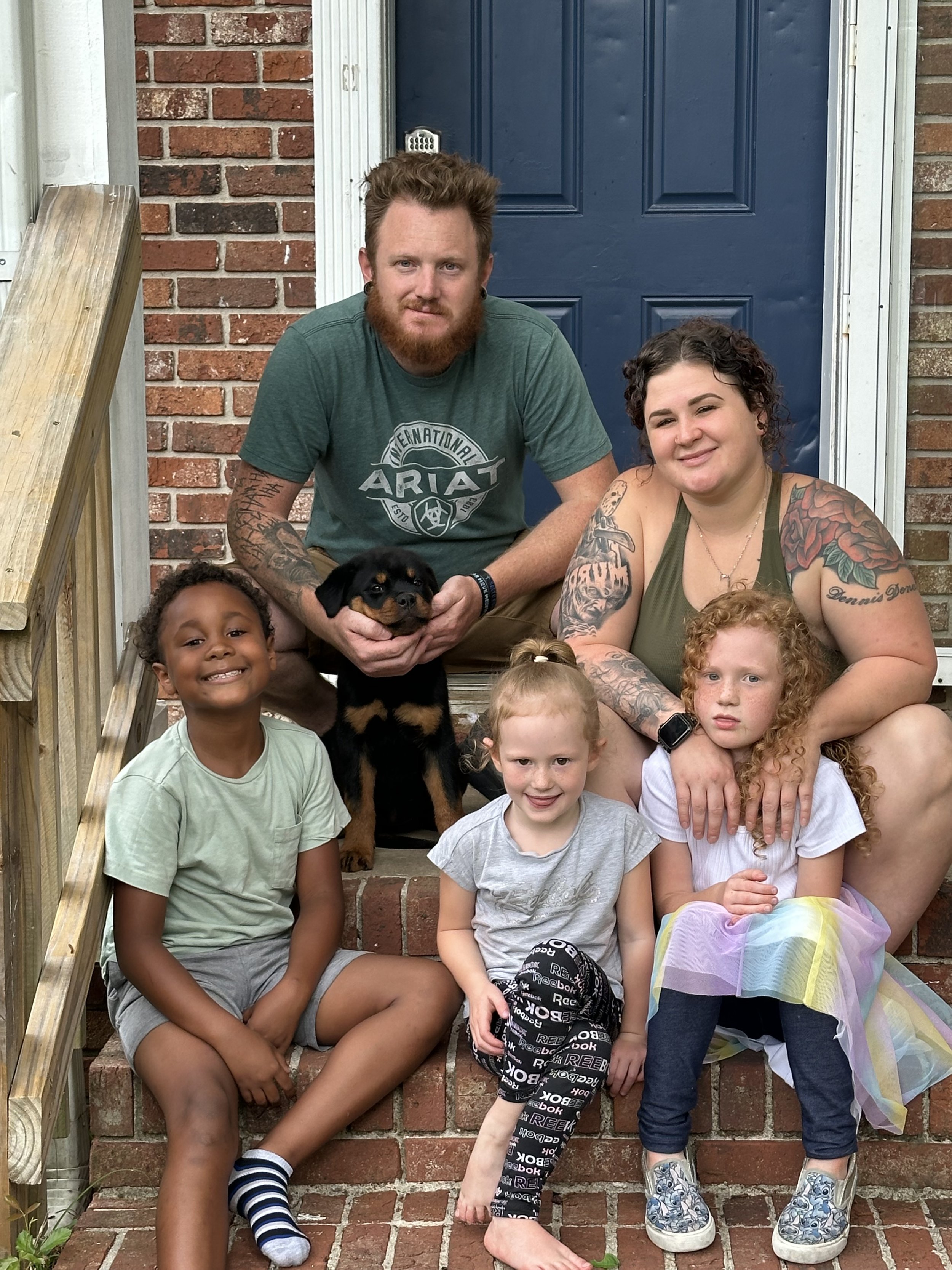A family of five poses on the front steps of their house with their dog. The group includes two adults and three children, all smiling and looking at the camera. The adults are sitting behind the kids with the dog, a small black and tan puppy, sitting on the steps between them. The background features a blue front door, a brick house, and a wooden staircase.