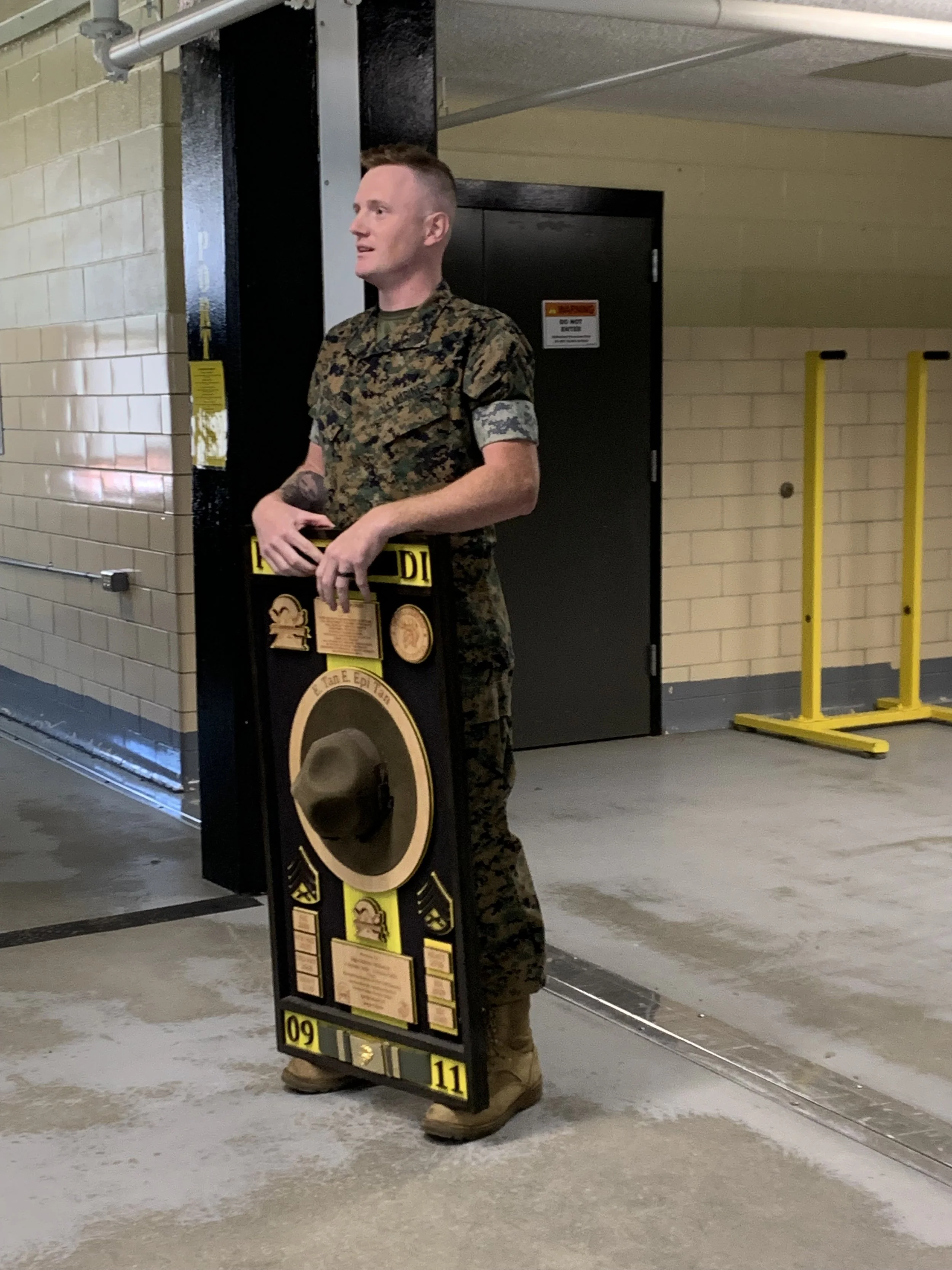 Man in camouflage uniform standing indoors, holding a large vintage-style alarm clock with a hat hanging on it.