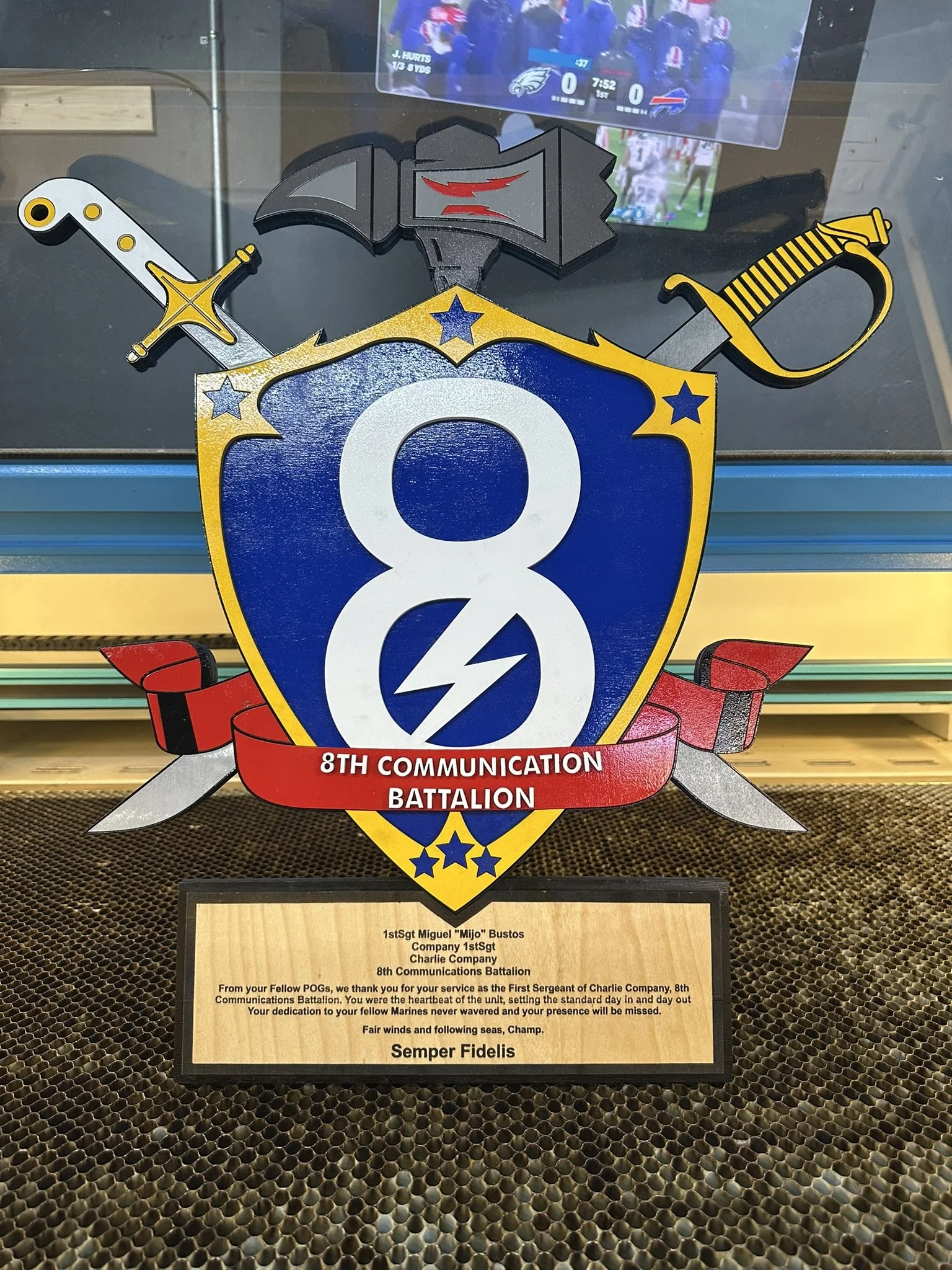 A plaque with a shield emblem for the 8th Communication Battalion. The shield features a large white number 8 with a lightning bolt, and is adorned with swords and a badge with a black and red flag. A red ribbon below reads "8TH COMMUNICATION BATTALION." Below the plaque, there is a wooden plaque with a dedication message.
