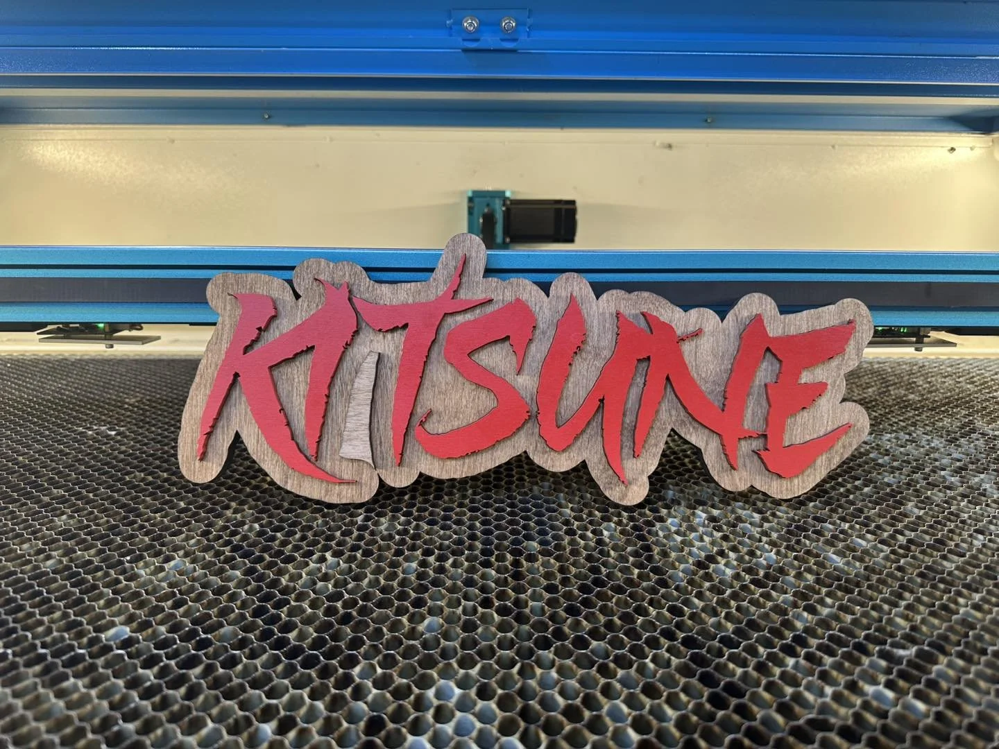 A wooden sign with the word 'KITSUNE' written in red graffiti style letters, positioned on a metal perforated surface with a metallic background and machinery.