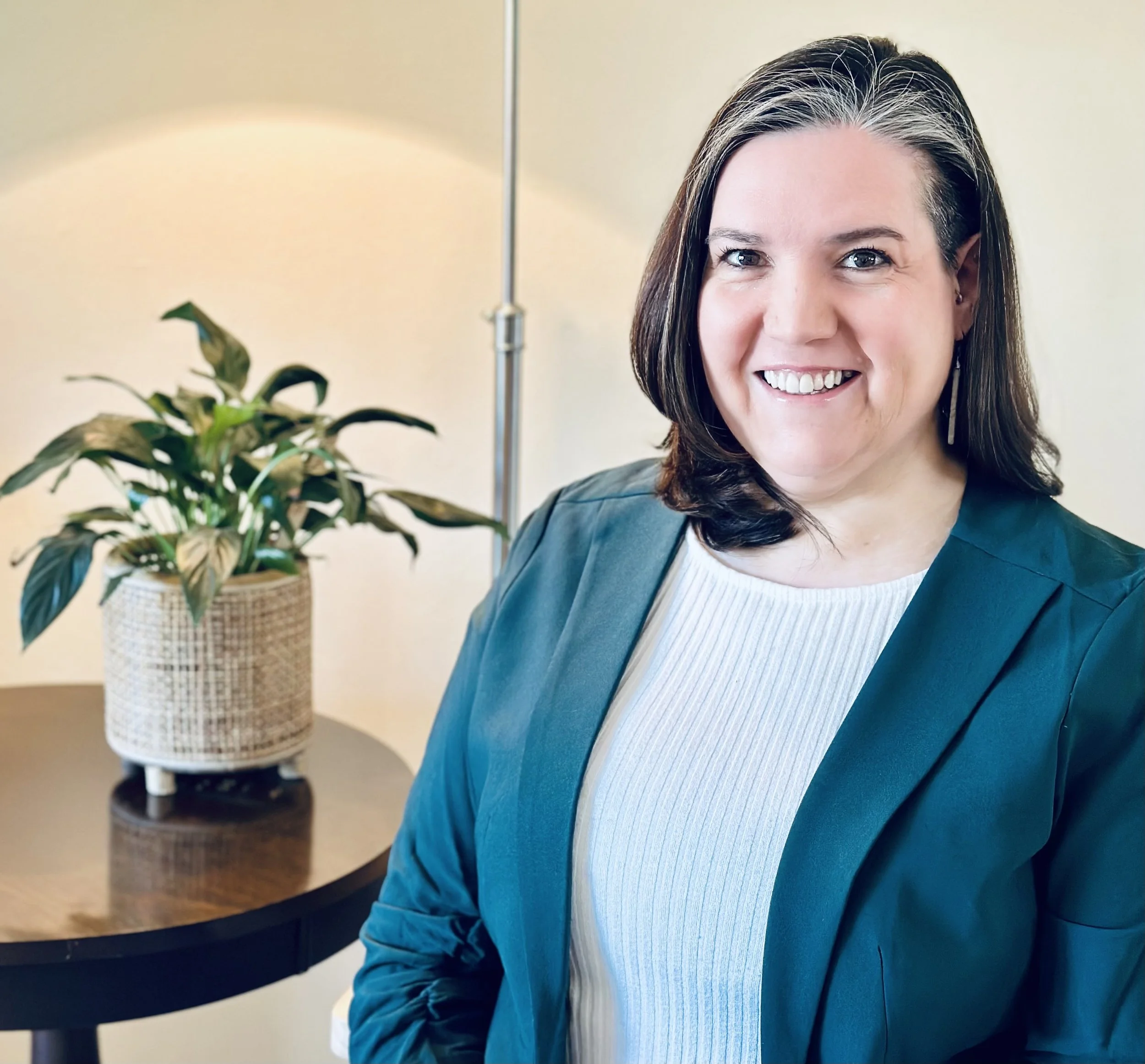 A smiling woman with shoulder-length dark hair and light streaks, wearing a teal blazer over a white ribbed top, standing in a room with a potted plant on a round wooden table behind her.