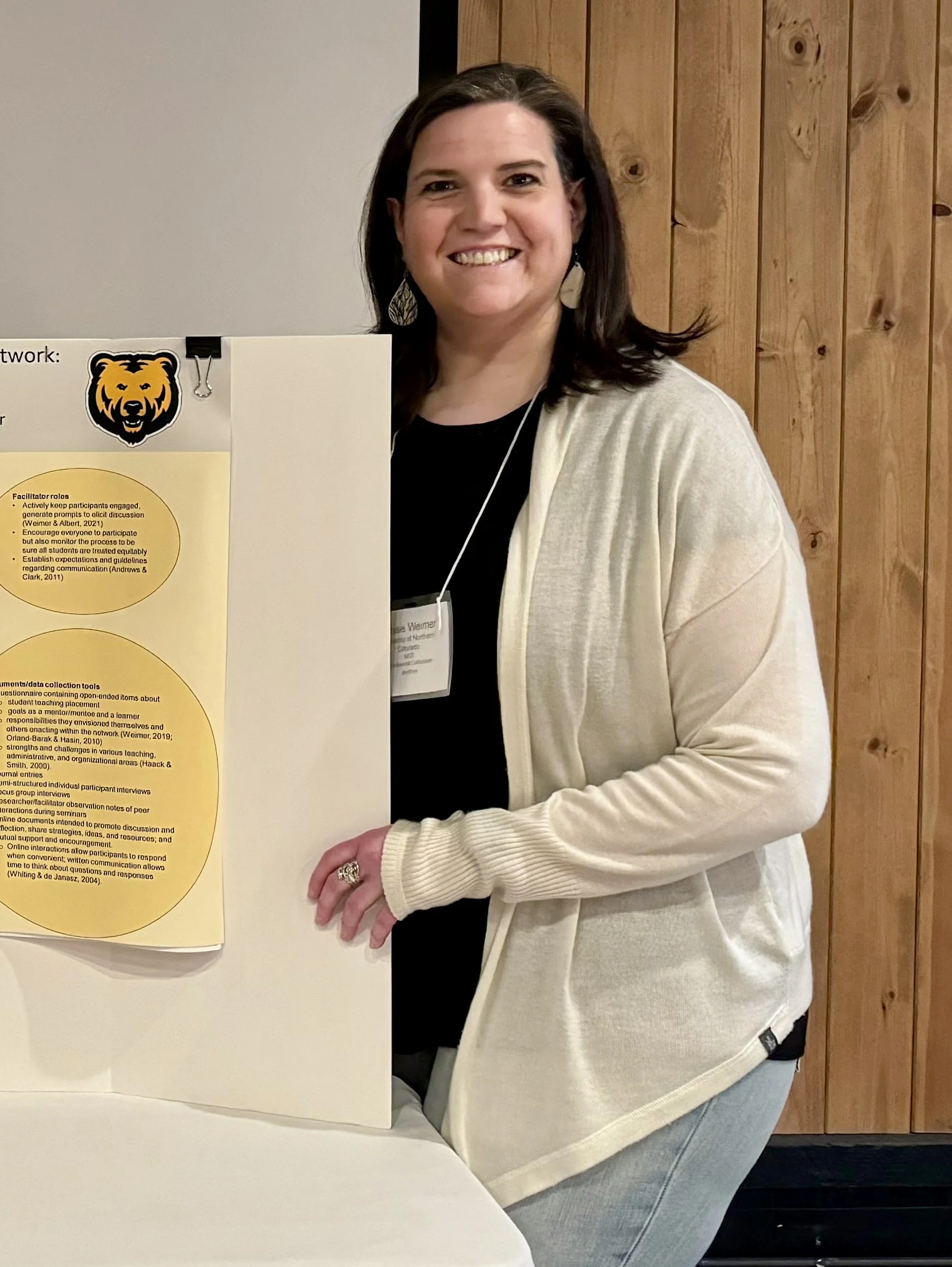 Woman with dark brown hair wearing a white cardigan and earrings standing next to research poster at conference.