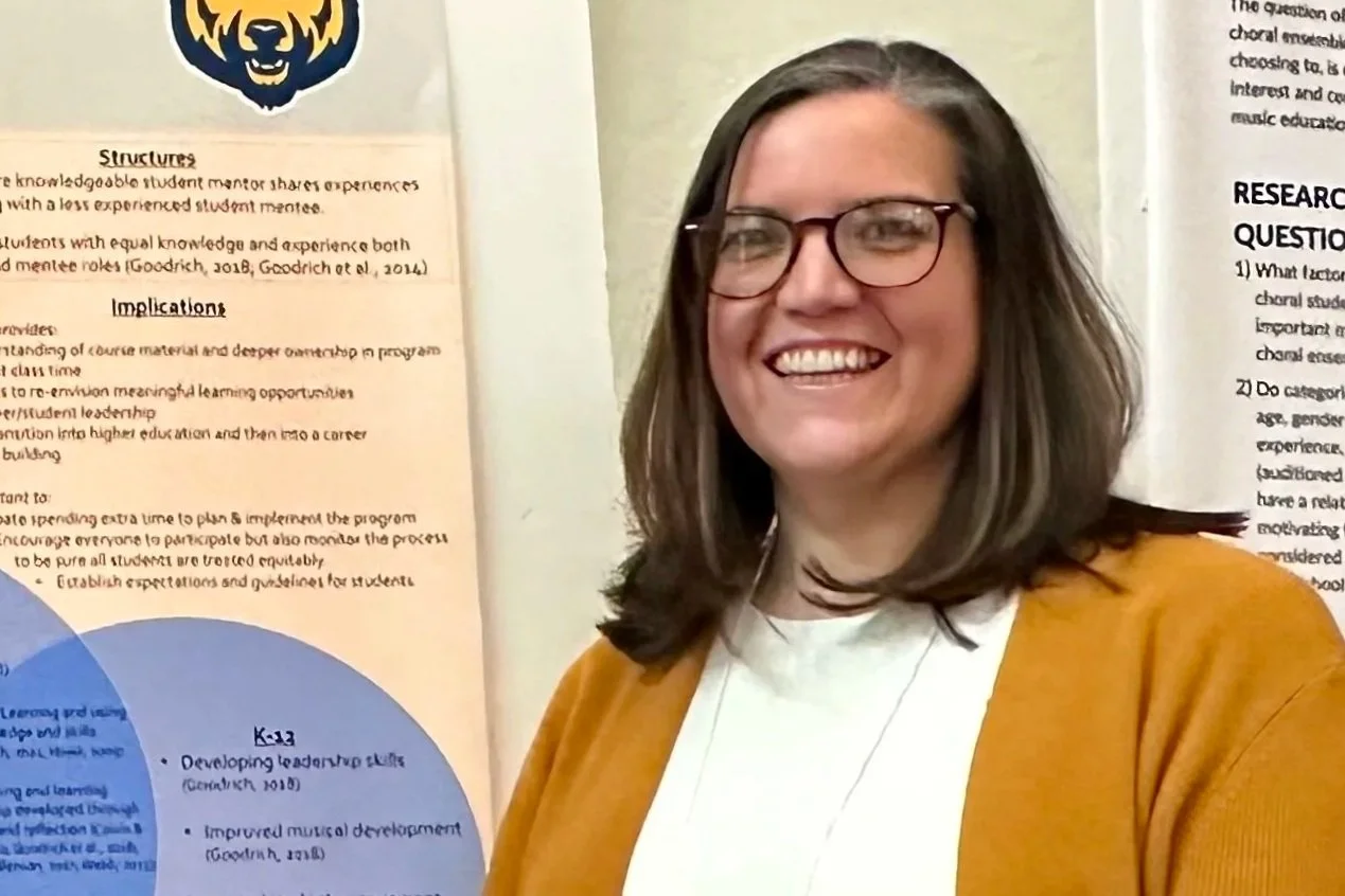 A woman with glasses smiling at a poster board discussing educational or research topics.