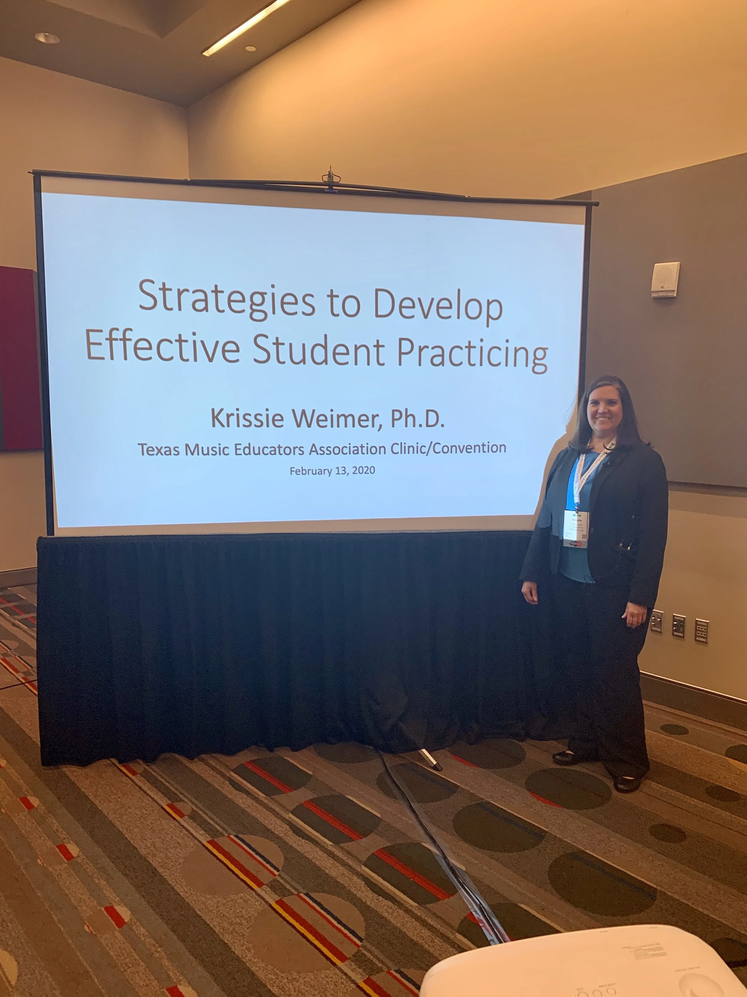 A woman standing next to a presentation screen that reads 'Strategies to Develop Effective Student Practicing' by Krissie Weimer, Ph.D., at the Texas Music Educators Association Clinic/Convention on February 13, 2020.