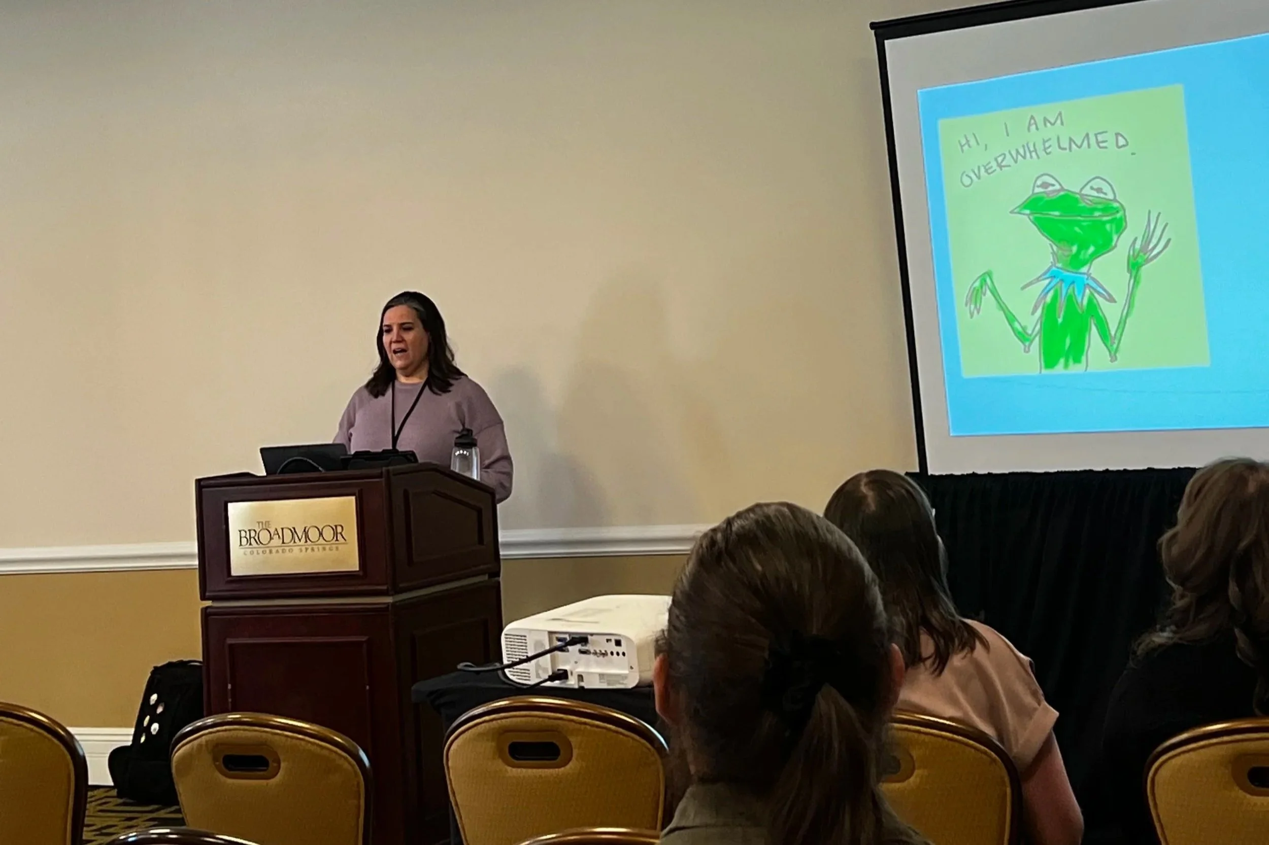 A woman giving a presentation at The Broadmoor hotel in Colorado Springs, standing behind a podium. A slide with a cartoon frog and the text 'Hi, I am overwhelmed' is projected on a screen. Audience members are seated facing the speaker.
