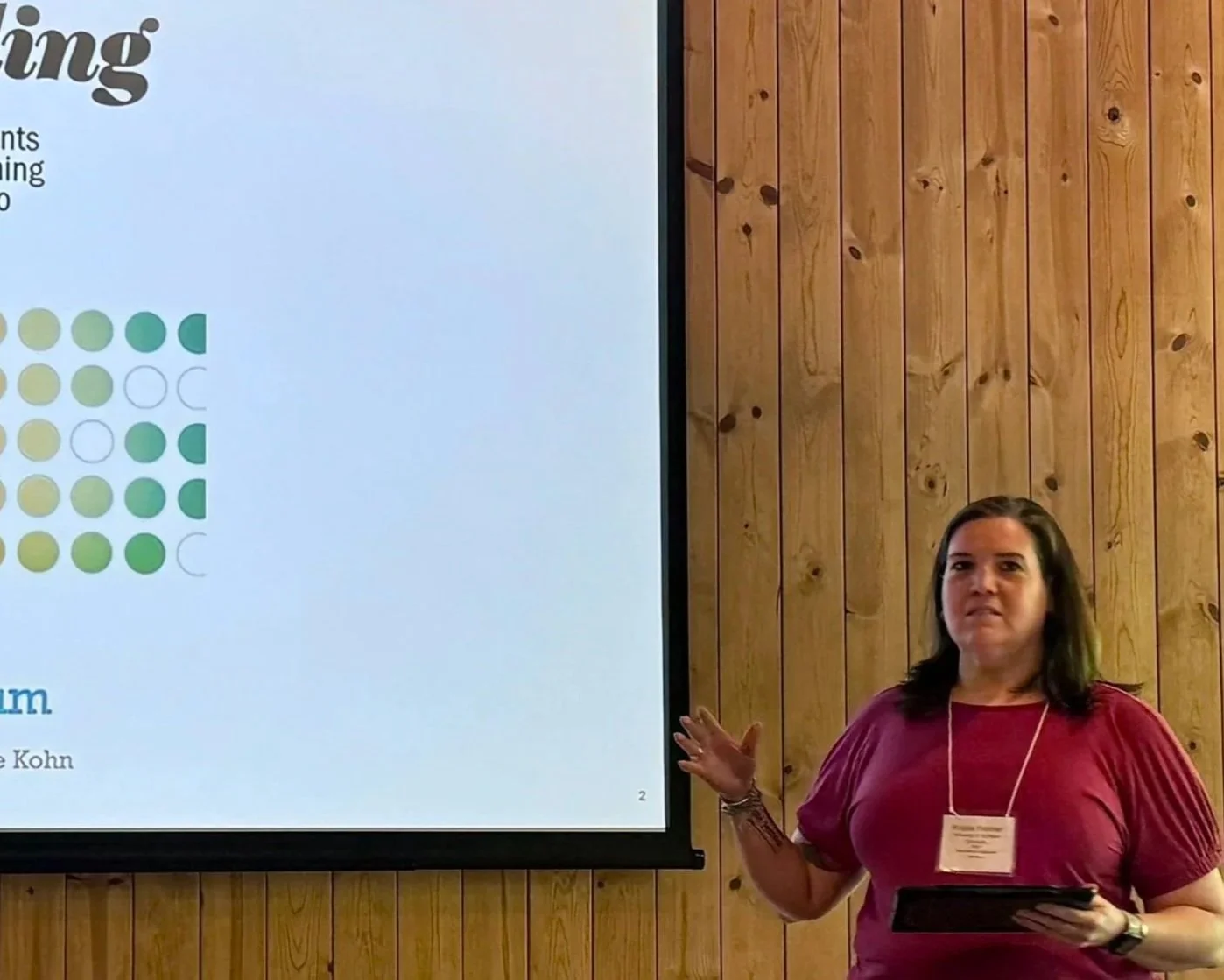 A woman giving a presentation next to a large screen with a slide that contains text, a graphic of circles, and some partially visible words. The background is a wooden wall.