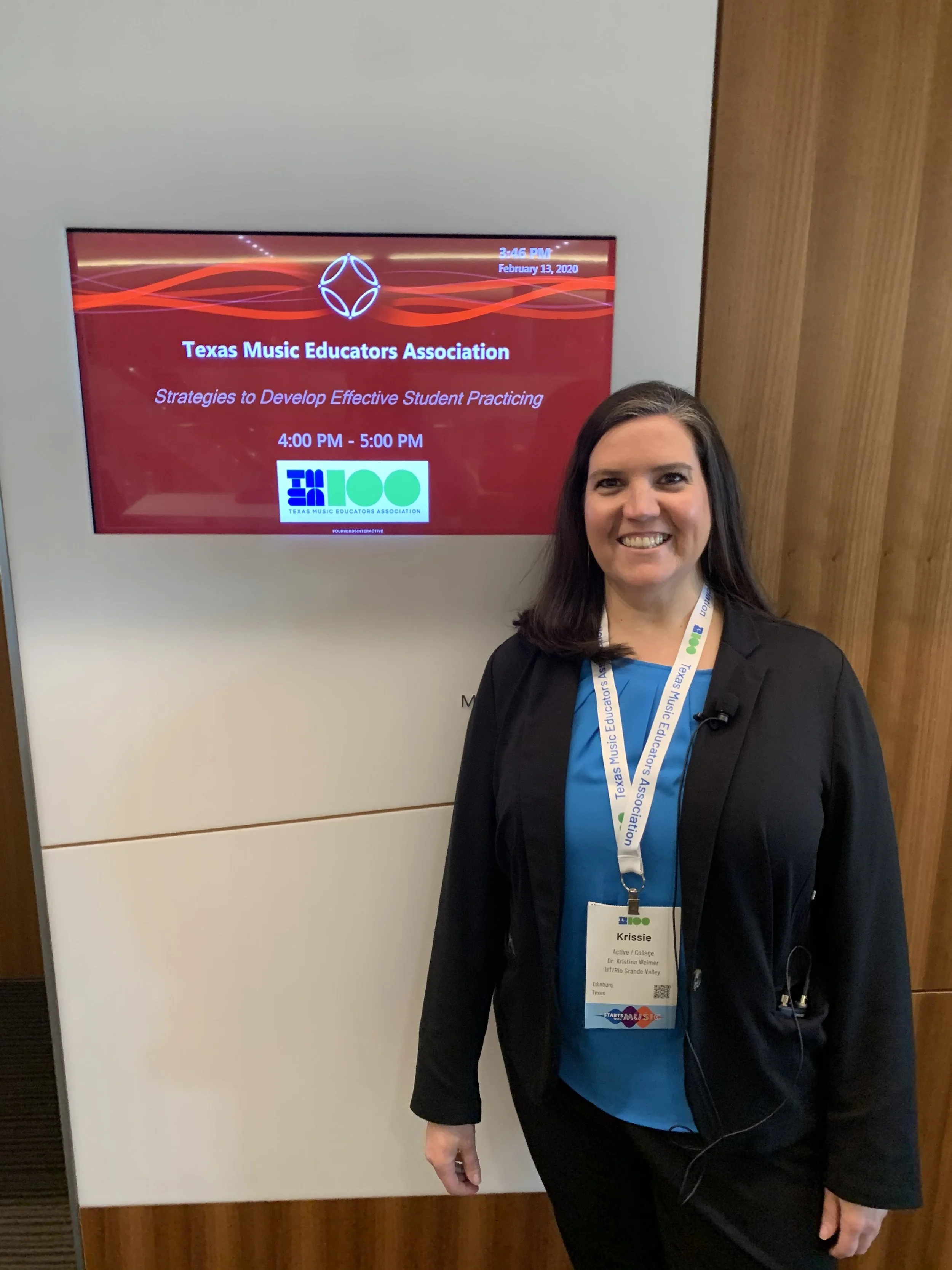 A woman smiling and standing in front of a digital display at a conference. The woman is wearing a black blazer, a bright blue shirt, and a conference badge with her name Krissie. The display shows the Texas Music Educators Association logo and information about a session on developing effective student practicing, scheduled from 4:00 PM to 5:00 PM on February 13, 2020.