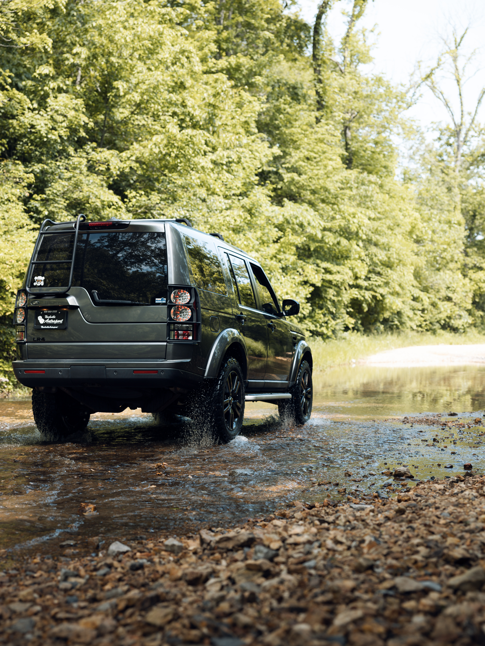 A black SUV driving through shallow water in a wooded area surrounded by green trees.