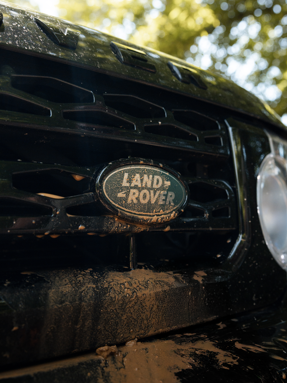 Close-up of a Land Rover vehicle with mud splattered on the grille and bumper, taken outdoors with trees in the background.