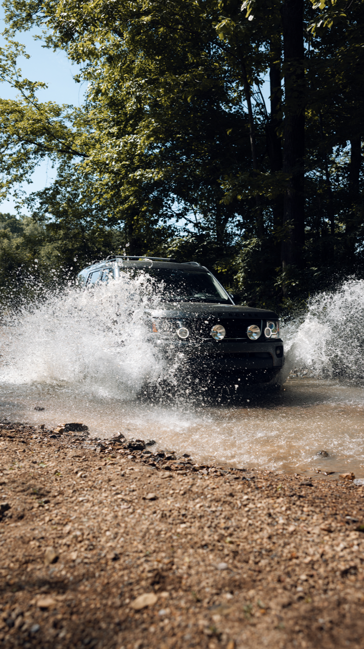 A black SUV driving through a shallow water crossing in a wooded area, creating a splash.