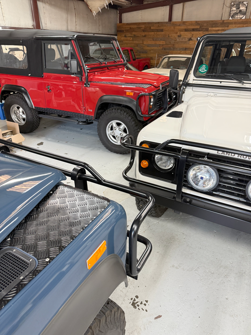 Multiple Land Rover vehicles parked inside a garage, including a red and a white model, with one blue vehicle partially visible in the foreground.