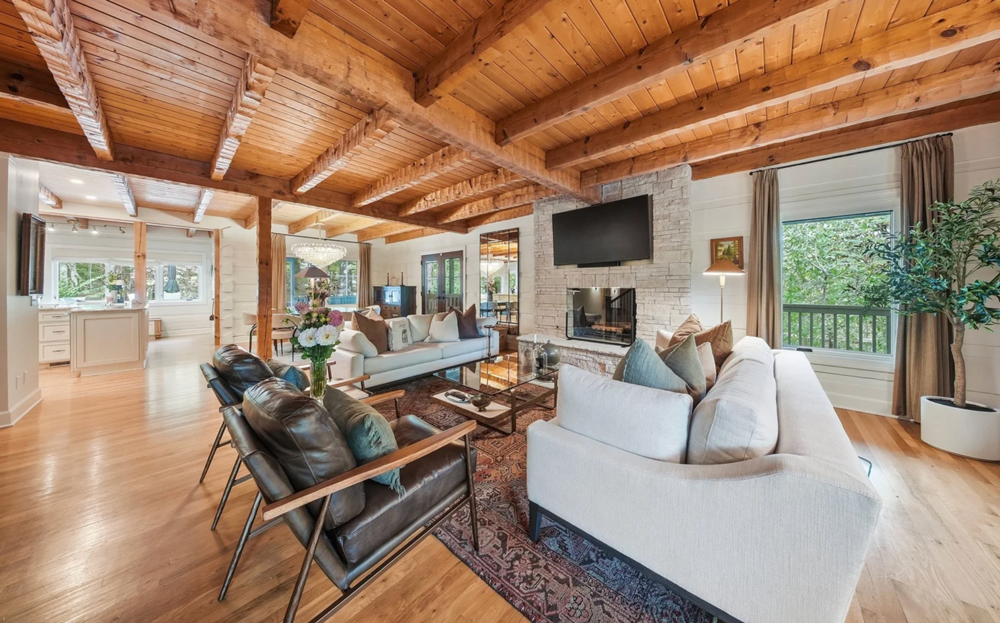 Spacious living room with wooden ceiling beams, white brick fireplace, flat-screen TV, large windows with curtains, and comfortable seating including two white sofas and leather armchairs.