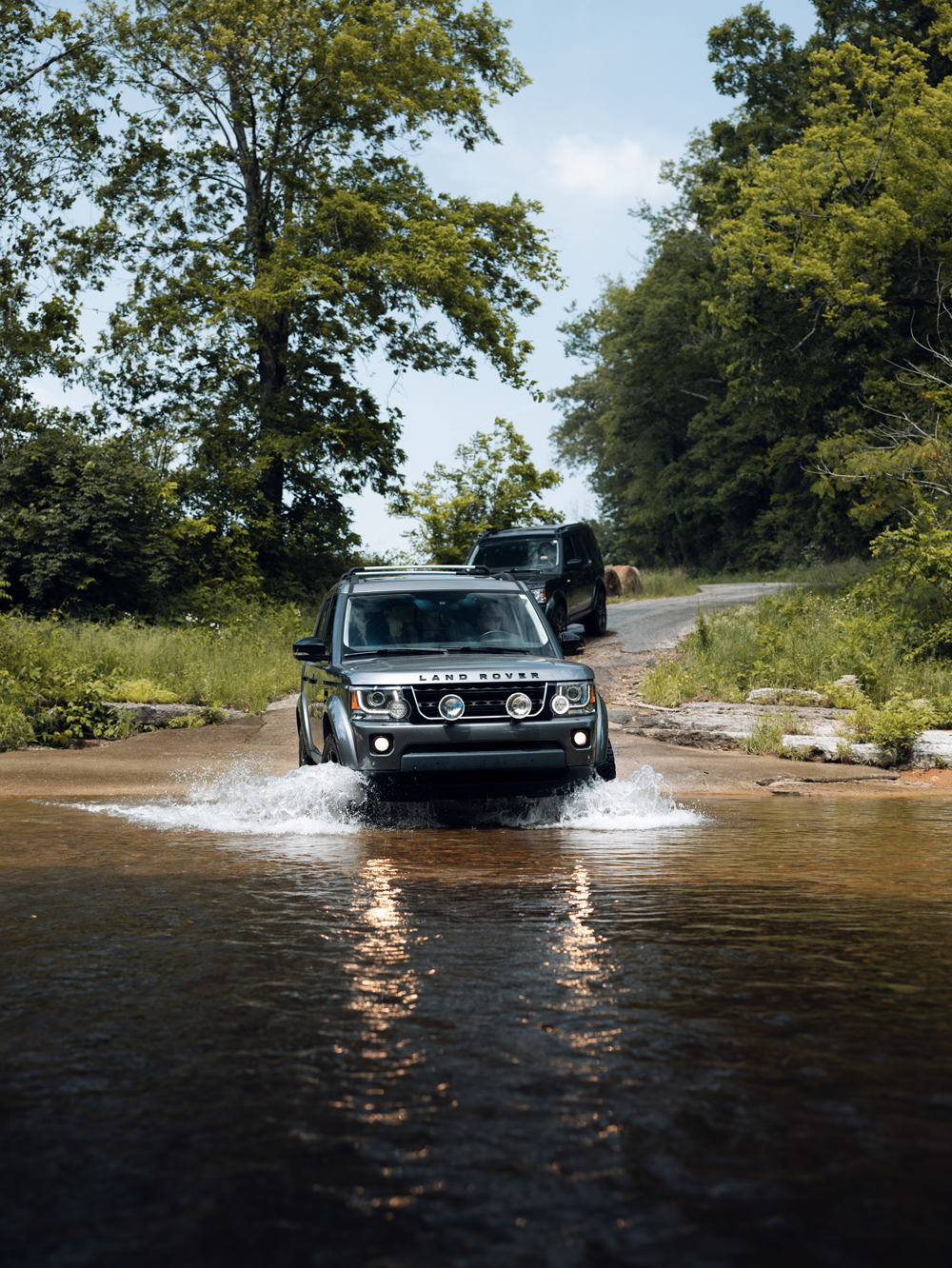 A Land Rover vehicle crossing a shallow river on a dirt trail surrounded by green trees in a forested area.