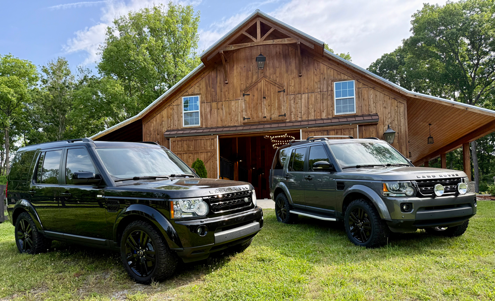 Two black Land Rover SUVs parked on grass in front of a large wooden barn with sliding doors, surrounded by green trees and a bright blue sky.