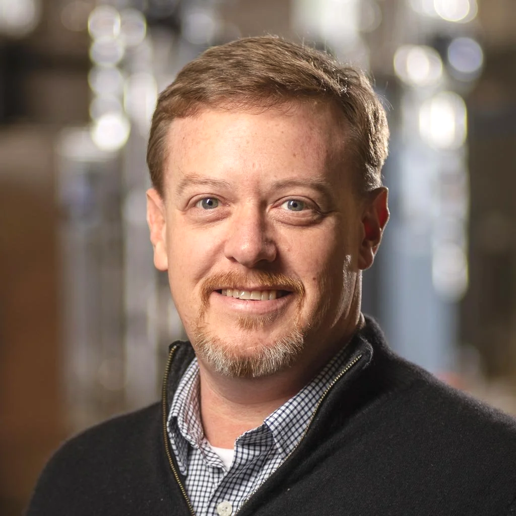 A smiling man with light brown hair, a mustache, and a goatee, wearing a checkered shirt and a black zip-up sweater, in an indoor setting with blurred background lights.