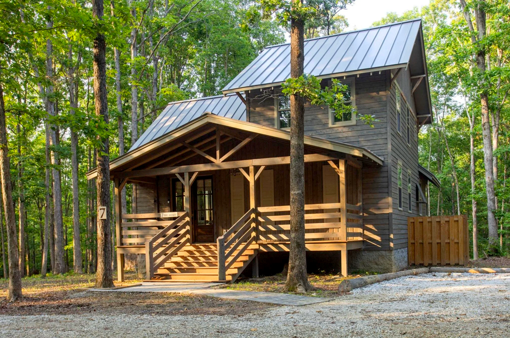 A two-story wooden house with a metal roof, surrounded by trees, features a front porch with wooden stairs and railings, set on a gravel lot.