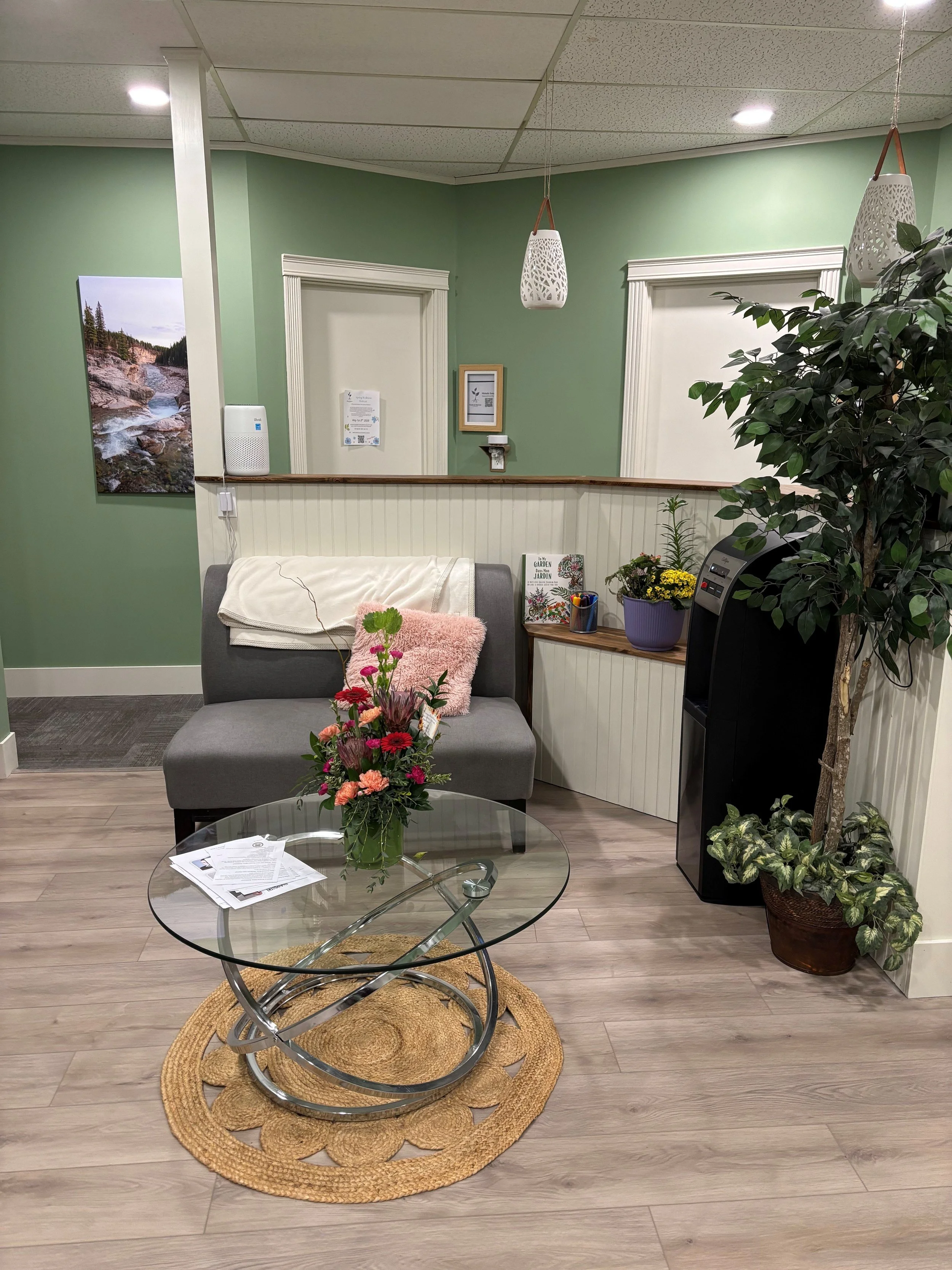 A waiting area with a glass coffee table on a woven rug, a gray upholstered chair with papers and a pink pillow, a flower arrangement, a white wall with framed pictures and a door, potted plants, and a water dispenser. The walls are painted light green, and the ceiling has recessed lighting.