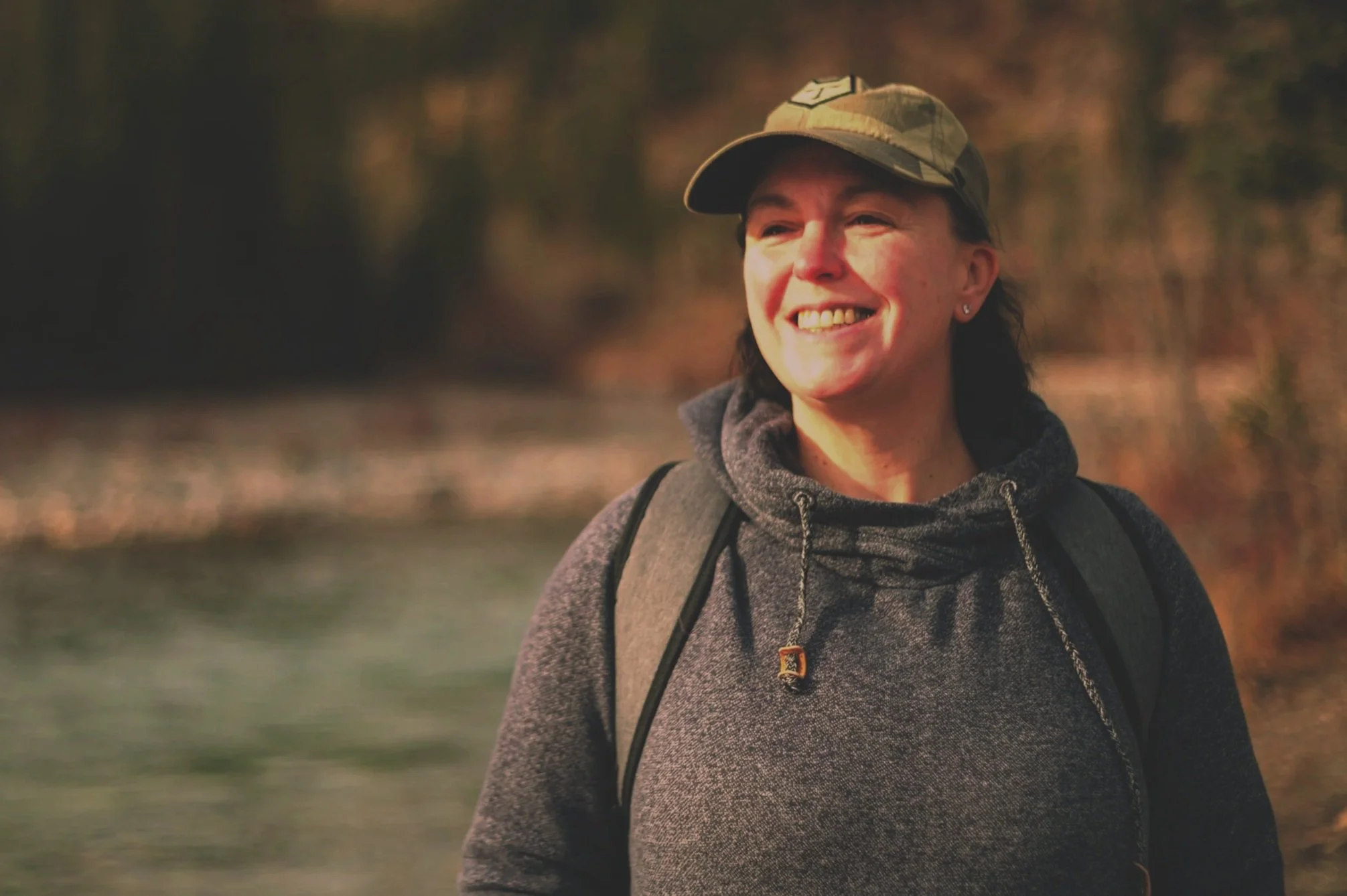 A woman smiling outdoors, wearing a camouflage cap, gray hoodie, and backpack, in a natural setting with blurred trees and water in the background.
