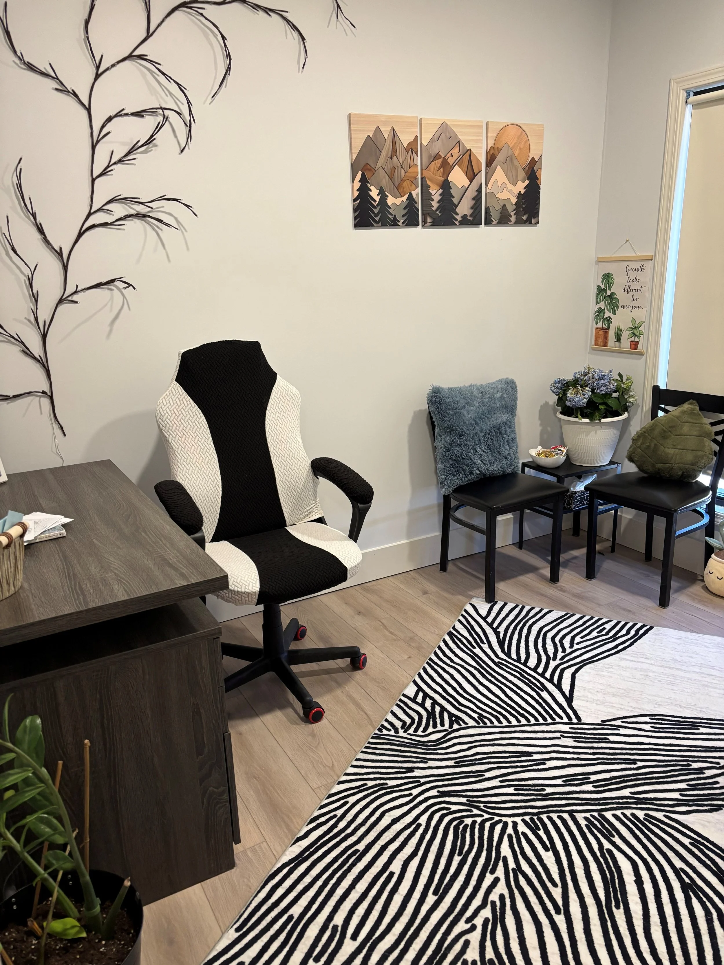 A therapy or counseling room with a black and white rug, a white and black ergonomic chair at a wooden desk, a small black table with a potted plant and snacks. Mountains and trees. There is a wall hanging with plants and a quote. Pillows and blanket