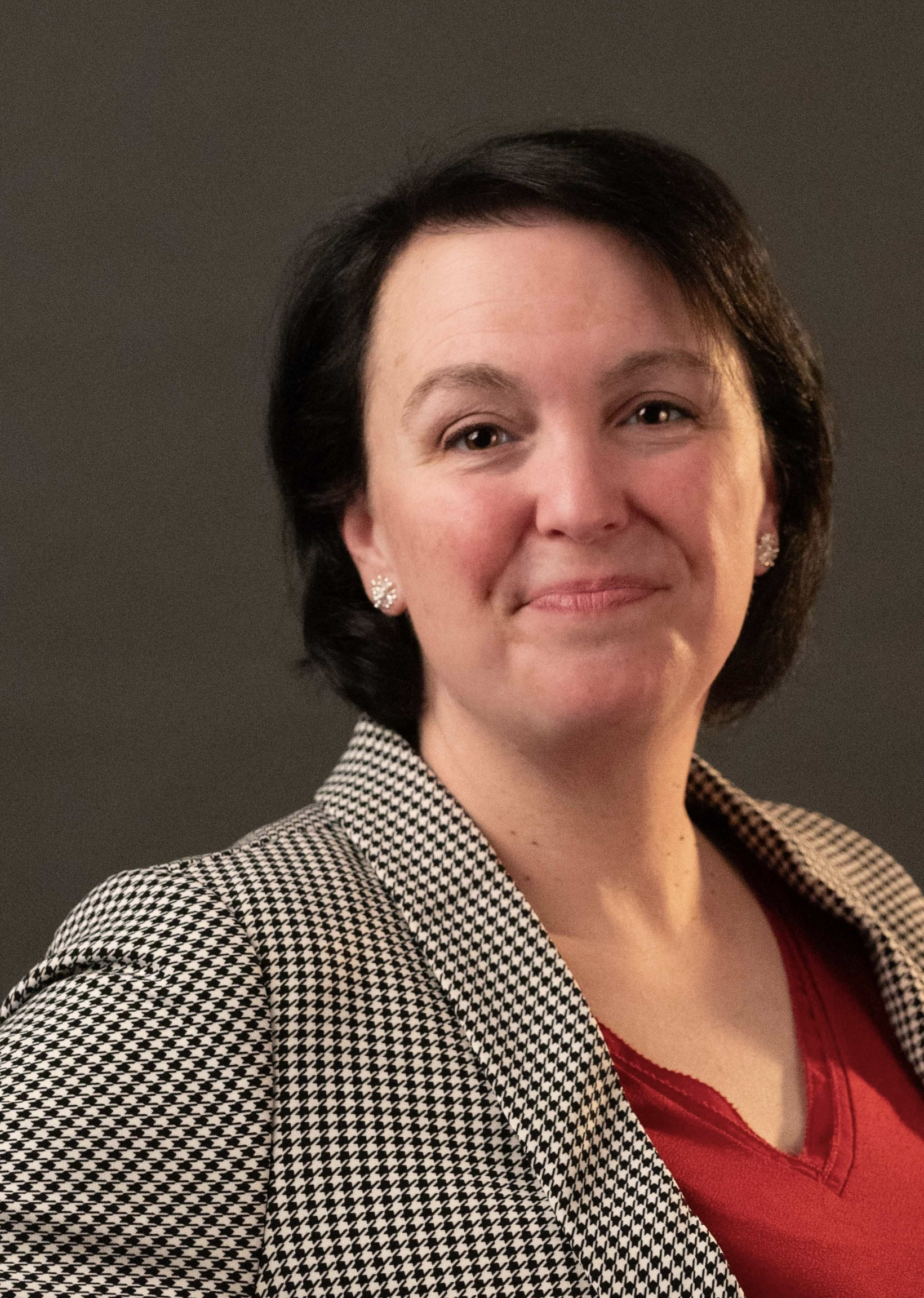 A woman with short dark hair, wearing earrings, a red top, and a black and white houndstooth blazer, smiling softly against a dark background.